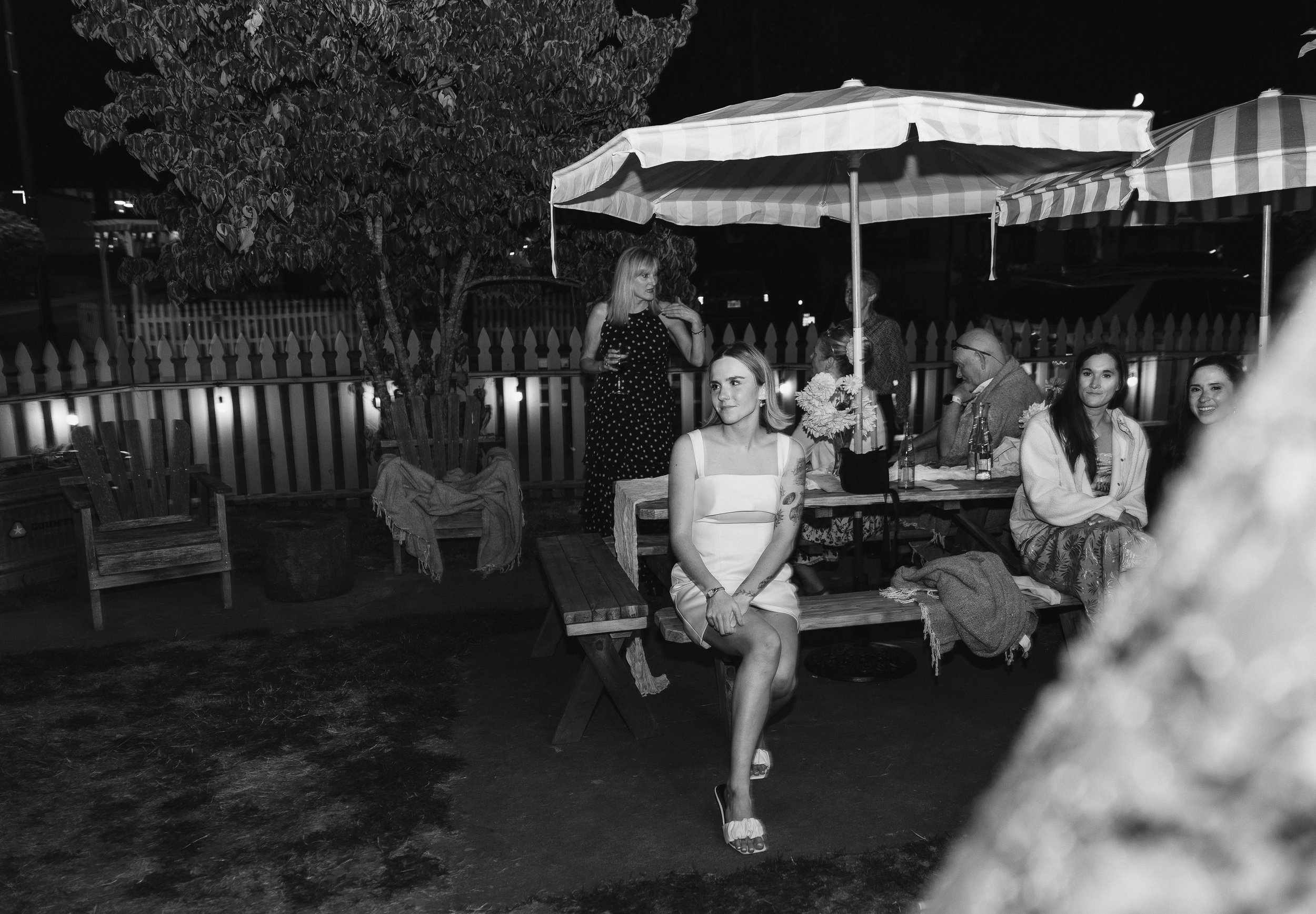 Nighttime outdoor gathering of five women and one man seated at a table with umbrellas, with one woman sitting on a bench and others chatting in the background, some holding drinks.