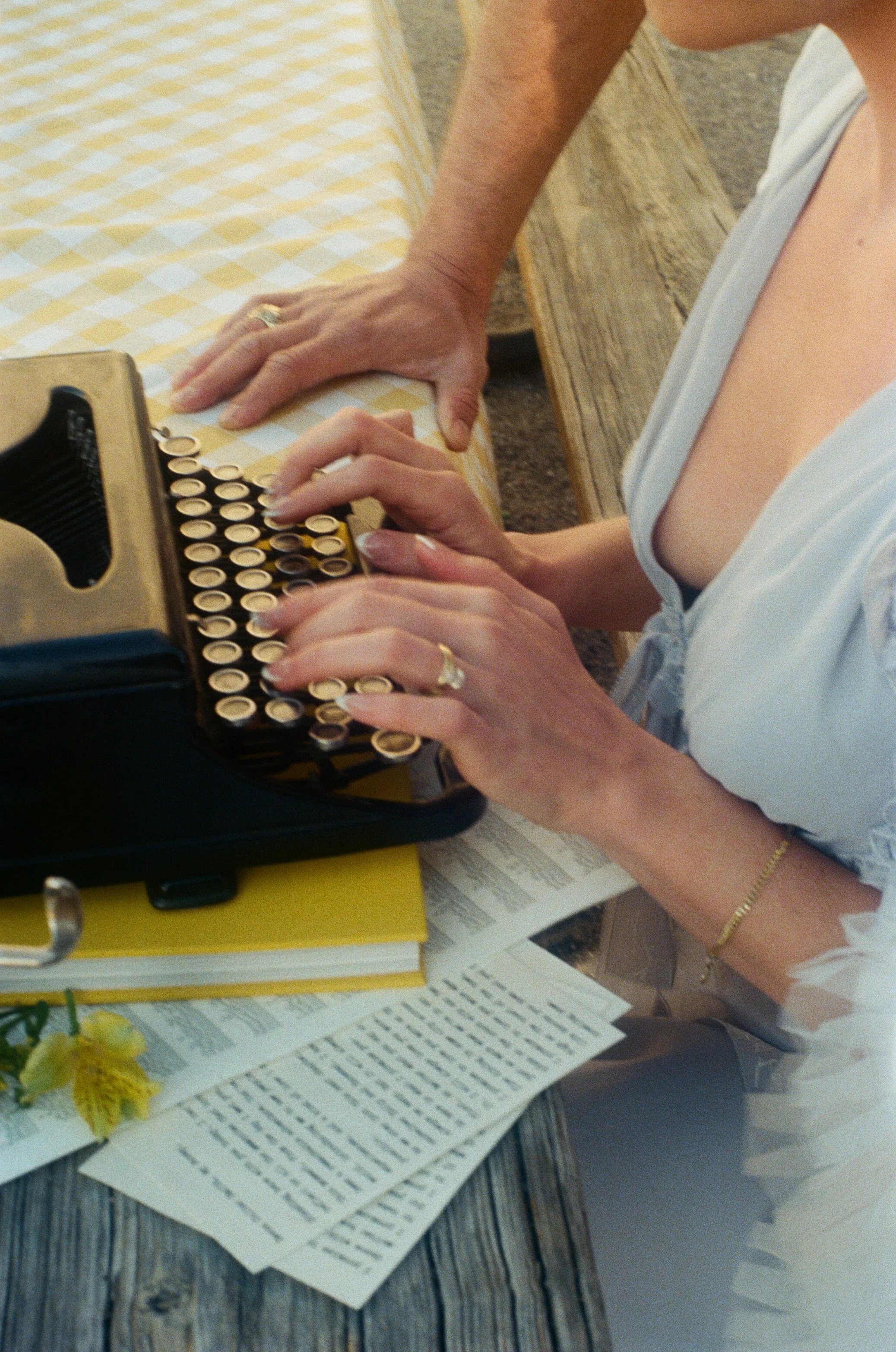 A woman types on a vintage typewriter at a wooden table, with a man leaning over to watch. There are papers, books, and a yellow flower on the table.
