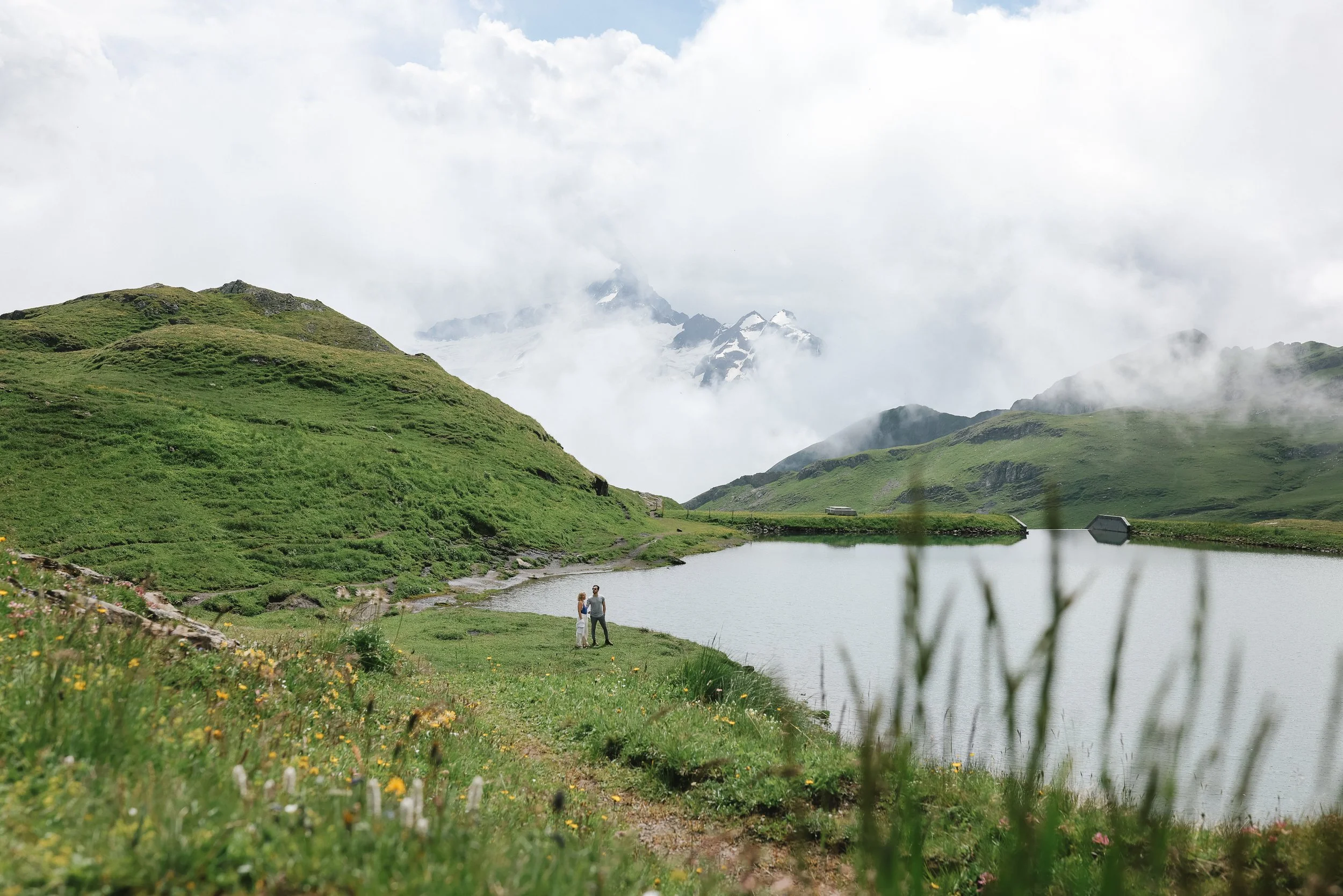 Couple standing by a lake surrounded by green hills and mountains with snow, partially clouded sky.