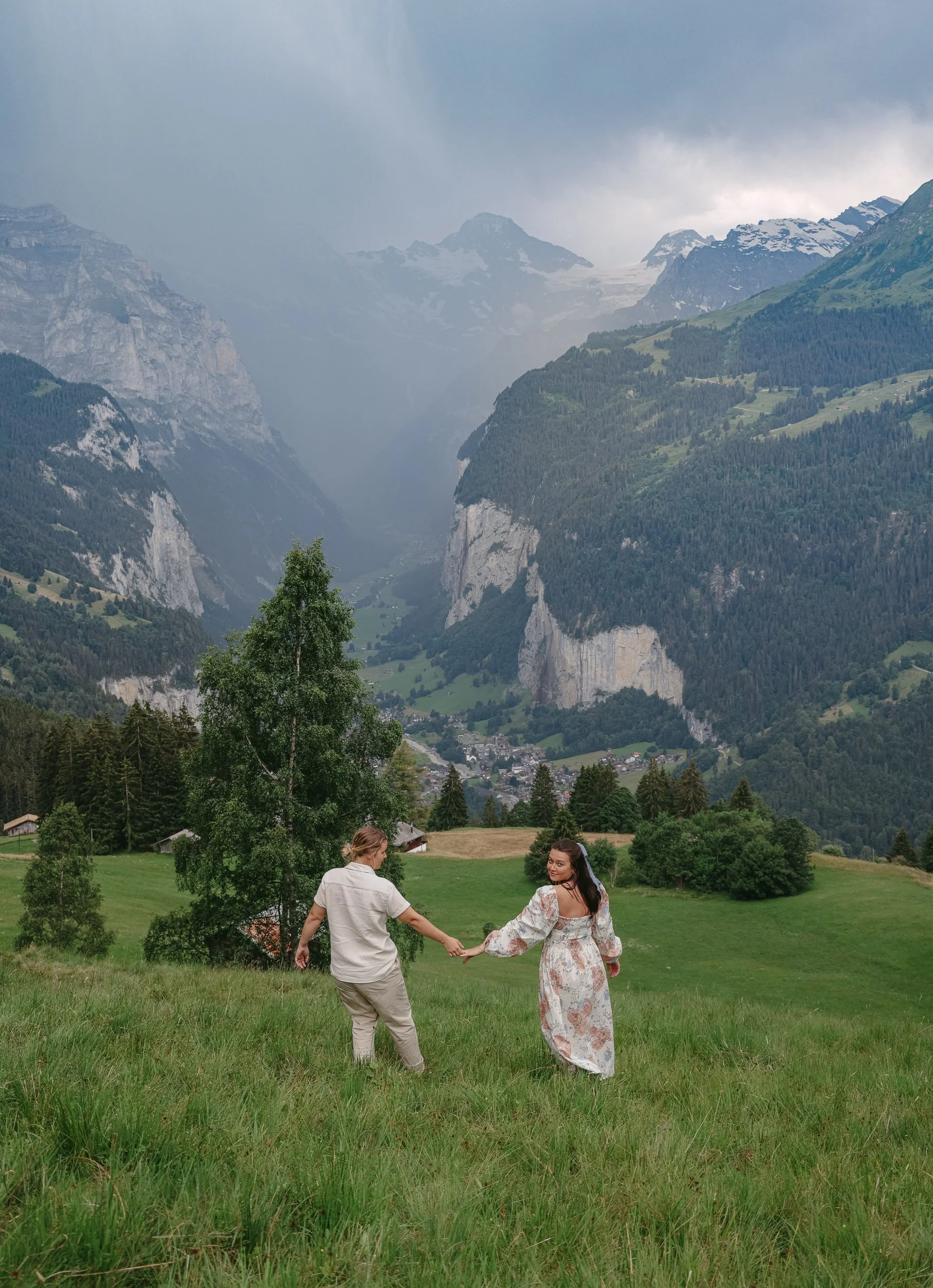 Two women holding hands and dancing in a green meadow with a mountain landscape in the background.