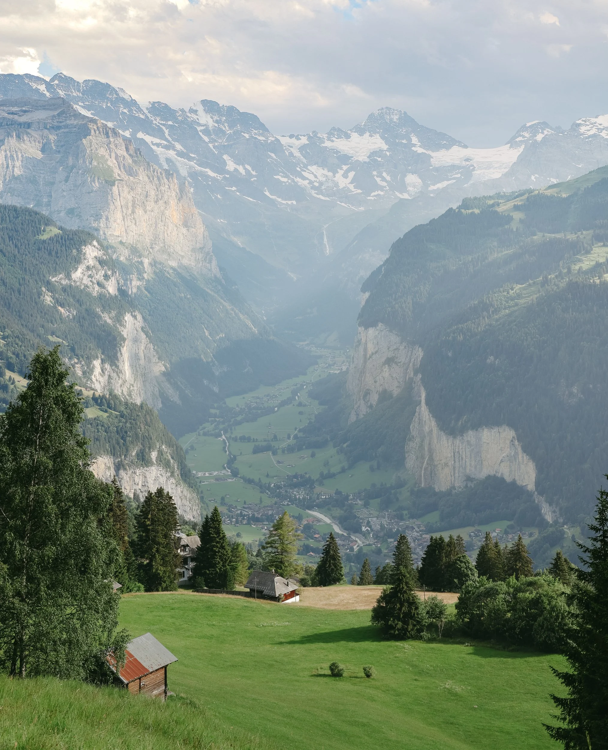 Scenic view of green valley with scattered houses, surrounded by tall evergreen trees and mountains in the background, with snow-capped peaks and cloudy sky.