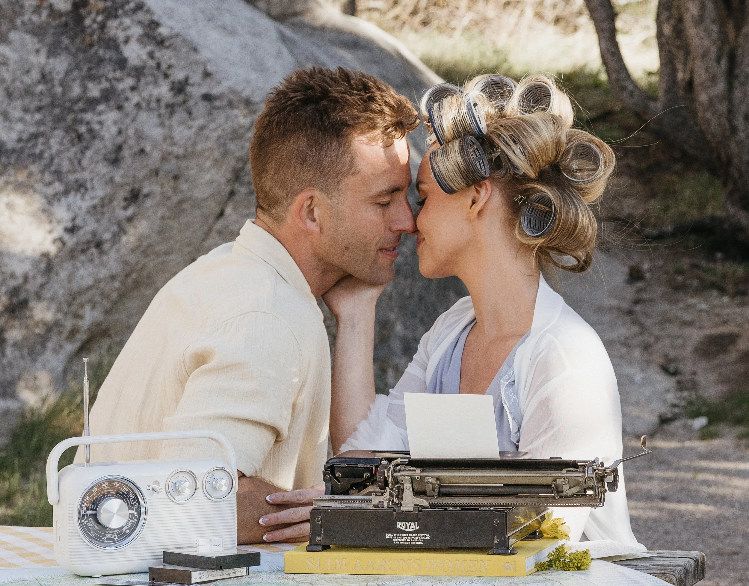 A couple with close noses touching, smiling, outdoors with rocks and trees, woman with curlers in hair, man in light shirt, vintage typewriter and radio on table.