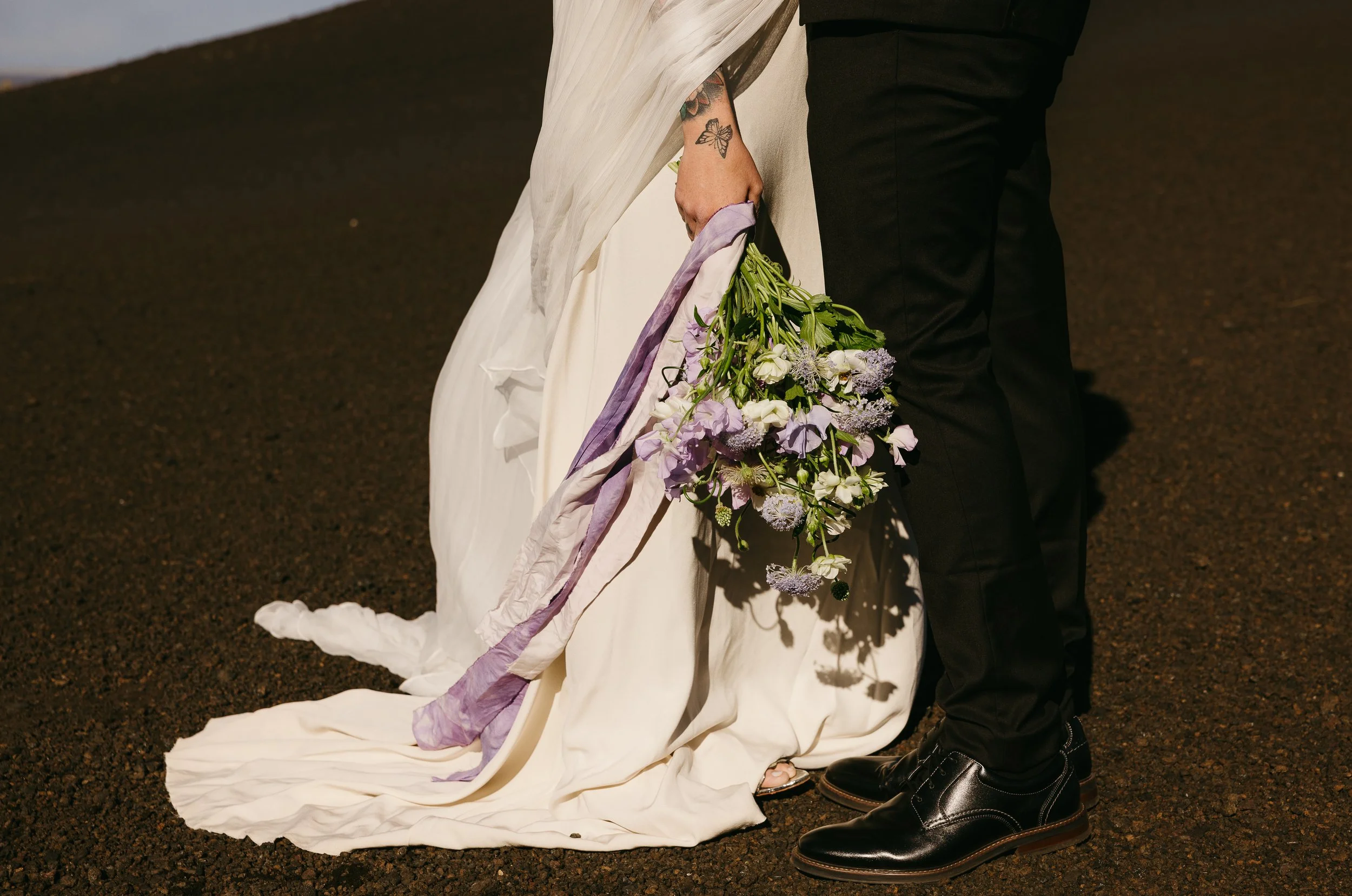 Close-up of a bride in a white wedding gown holding a bouquet of flowers, standing next to a groom in black pants and shiny black shoes on a dark street.