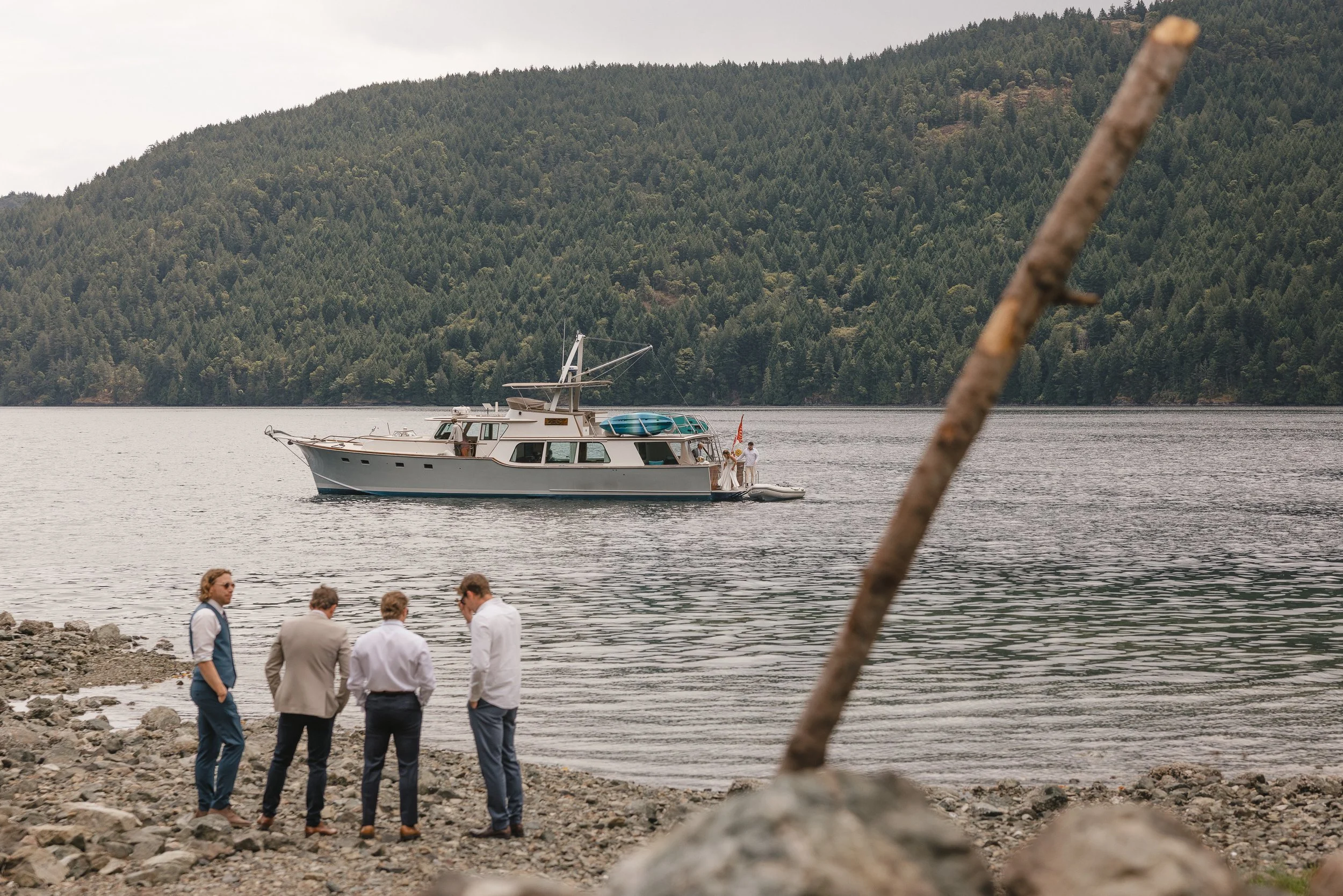 A group of five men dressed in formal and casual attire standing on a rocky shore, looking toward a yacht on the water with forested hills in the background.