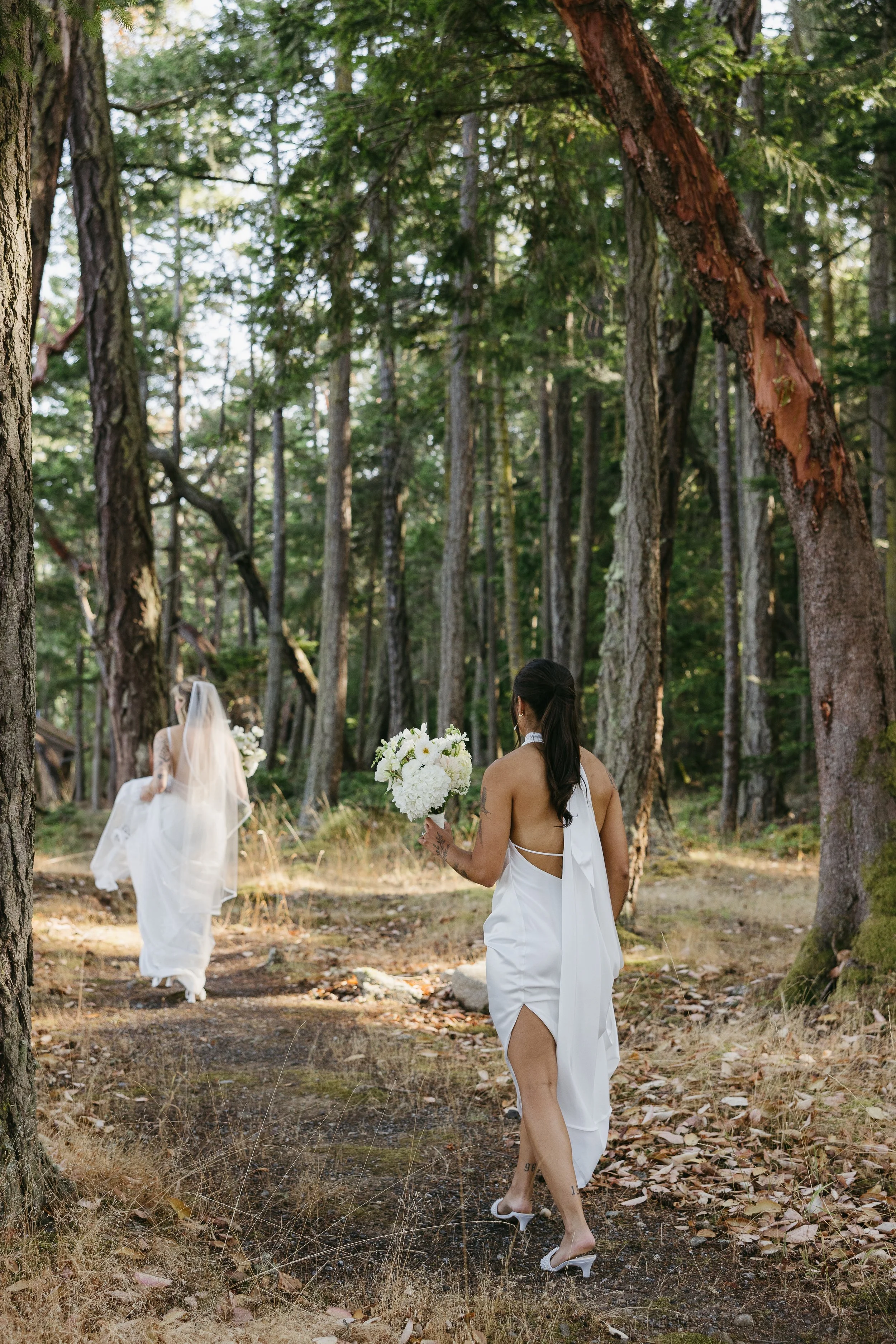 Two women in white dresses in a forest; one woman, with long black hair, holding a bouquet of white flowers, is walking towards another woman in a wedding dress with a veil.