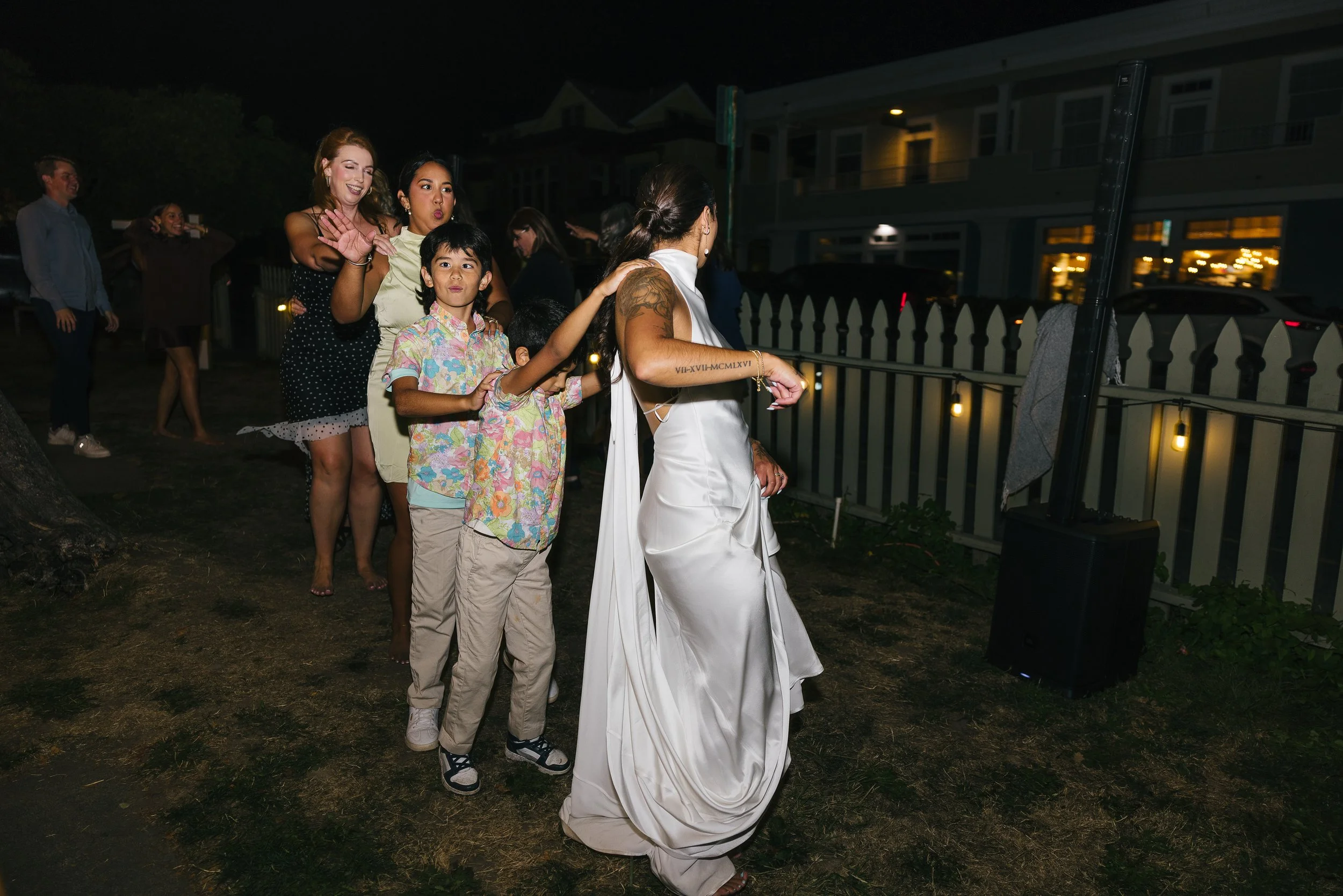 A woman in a white dress leading a line of people during a nighttime outdoor event, with a fence and houses illuminated in the background.