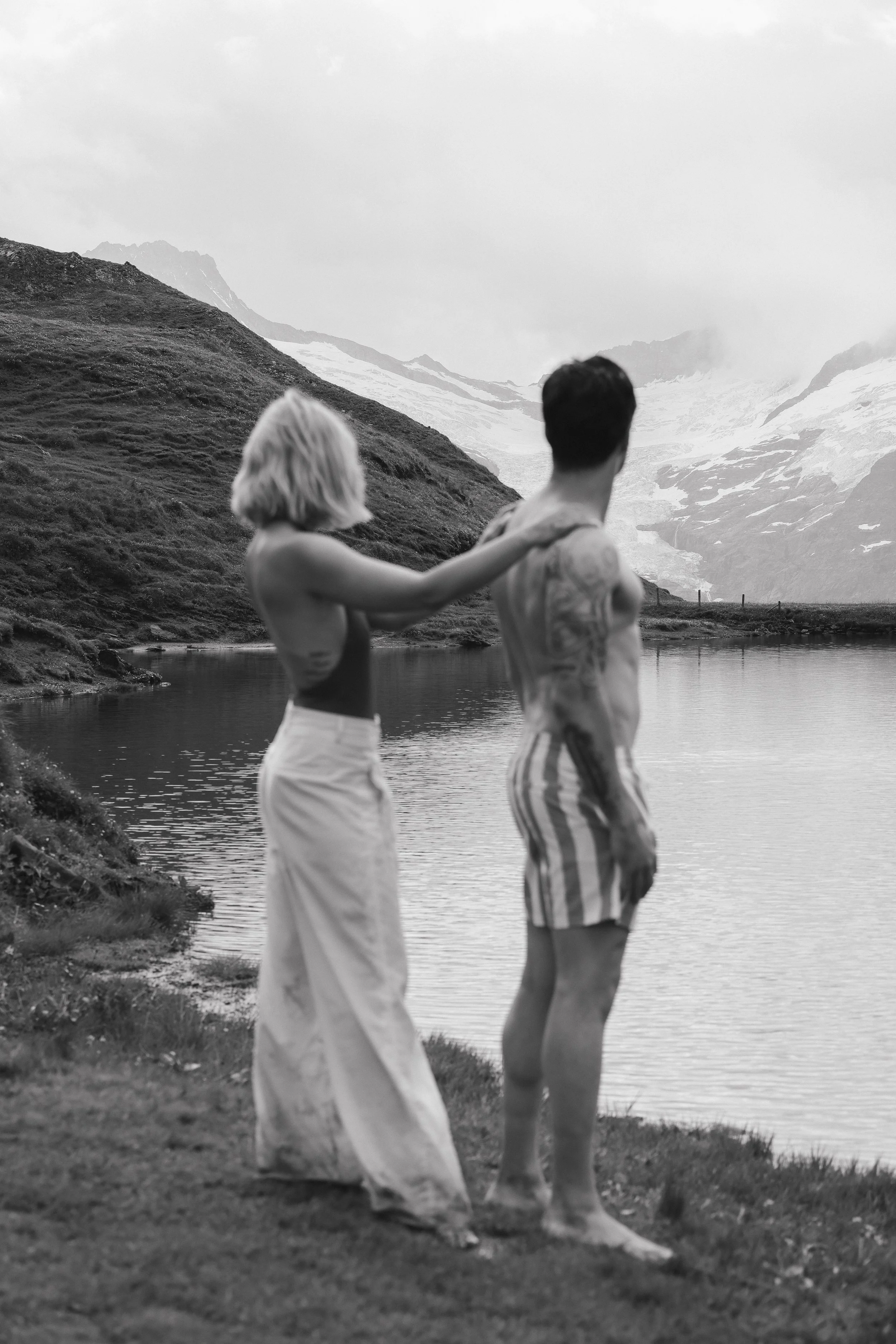 A black and white photo of a woman and man standing near a lake in a mountainous area, with the woman gently touching the man's shoulder as they face the water and mountains.
