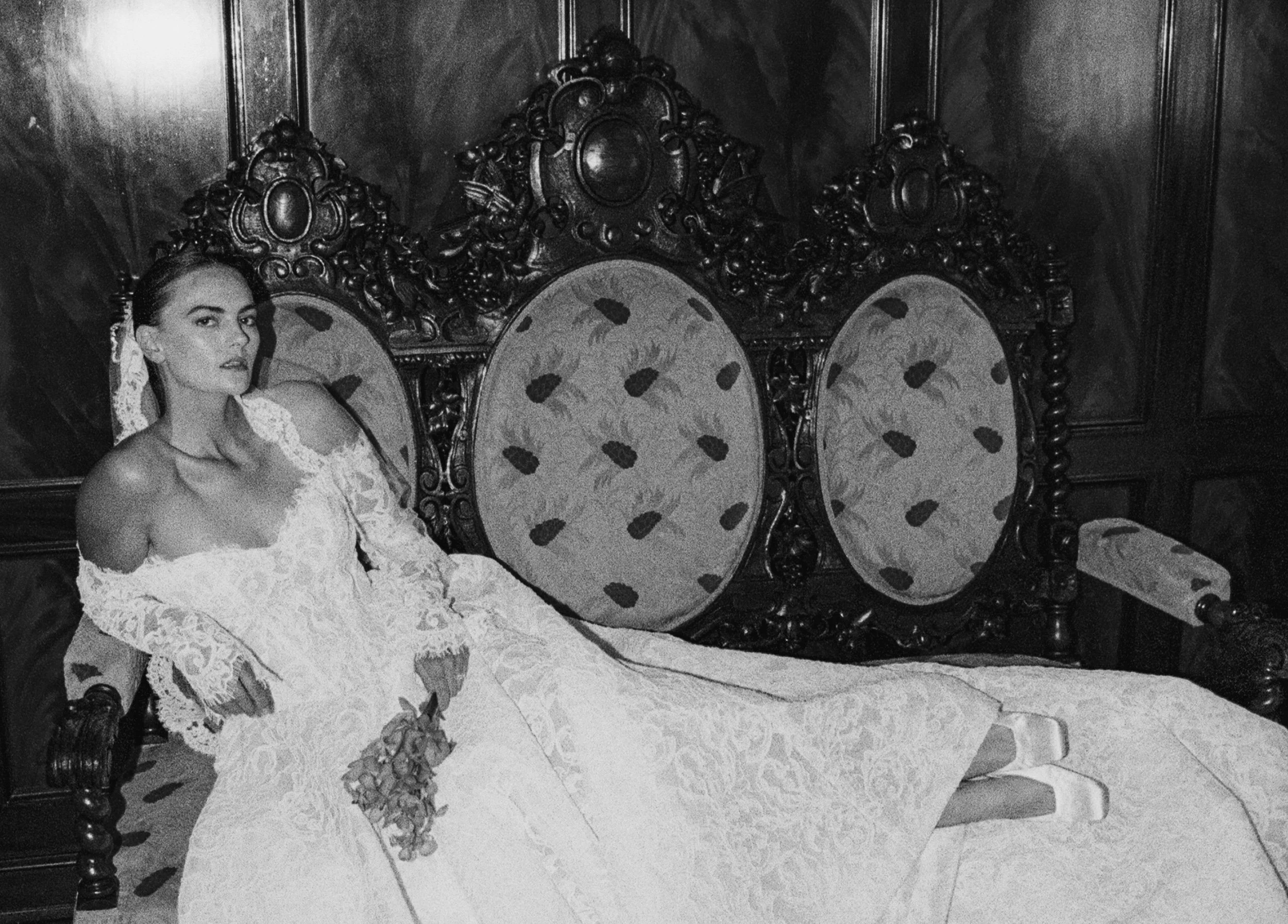 A woman in a lace wedding dress lying on an ornate wooden bed with tall, carved headboard and upholstered oval panels. She is holding a small bouquet of flowers and looking at the camera.
