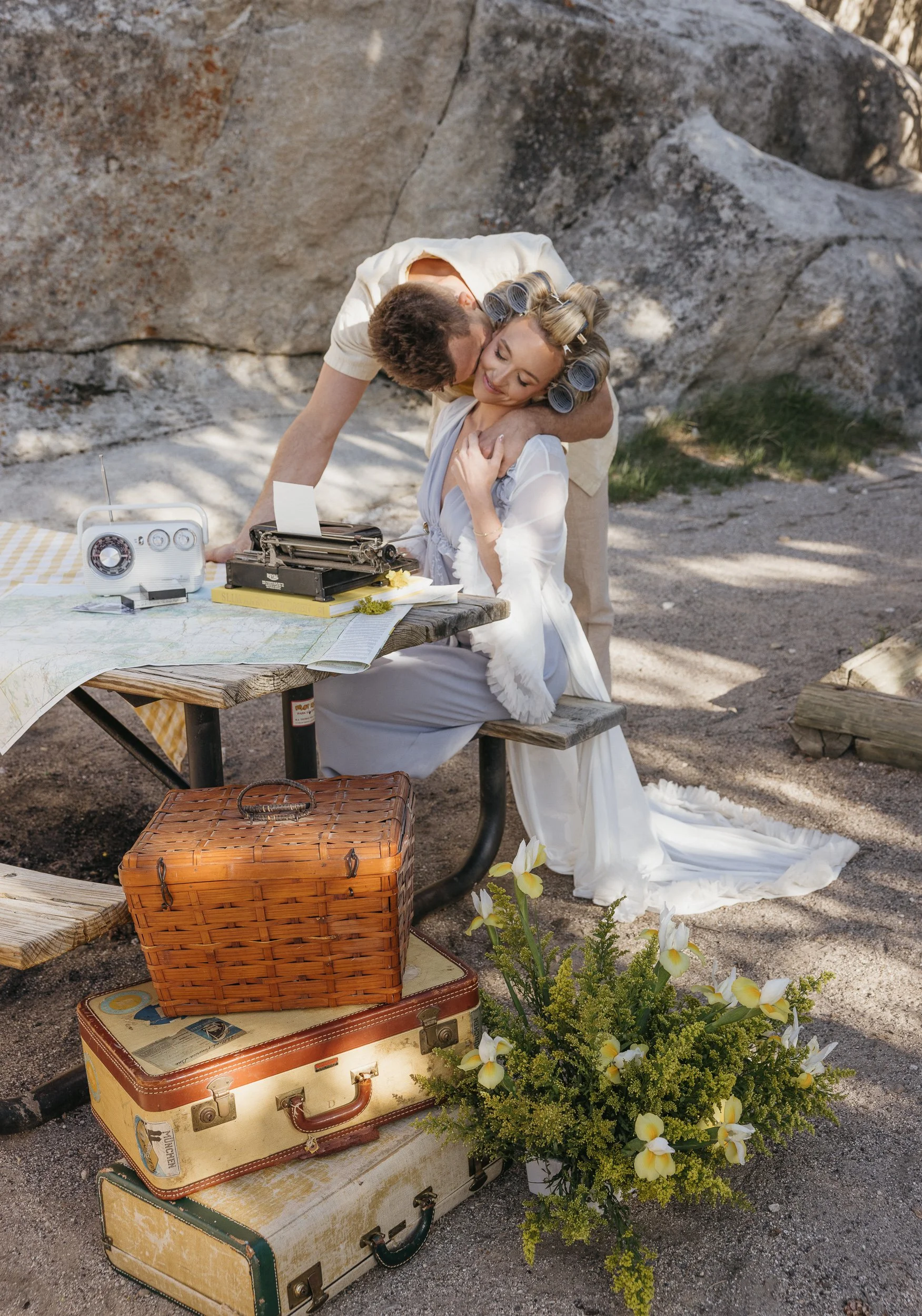 A man and a woman dressed in vintage clothing are outdoors, with the man leaning over and hugging the woman as she sits at a picnic table with a typewriter. The woman has her hair in curlers and is smiling, with a large bouquet of flowers placed on t