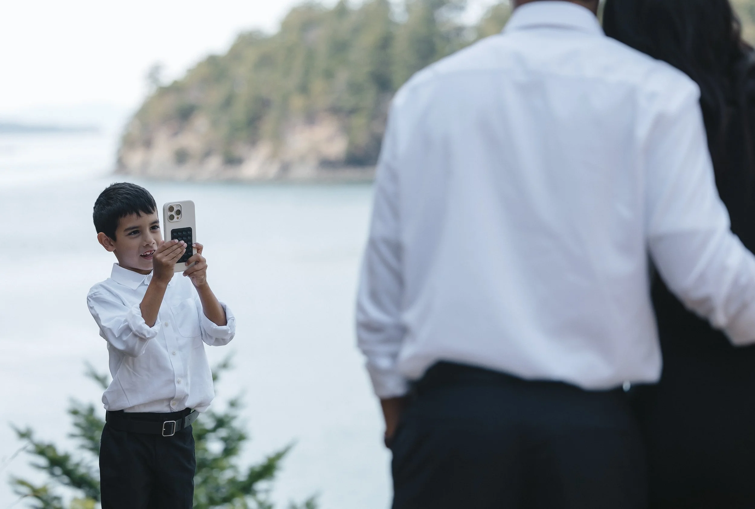 A young boy in a white shirt and black pants is taking a photo with a smartphone. Two adults are standing nearby with their backs to the camera, and a scenic outdoor background of water, trees, and hills is visible.