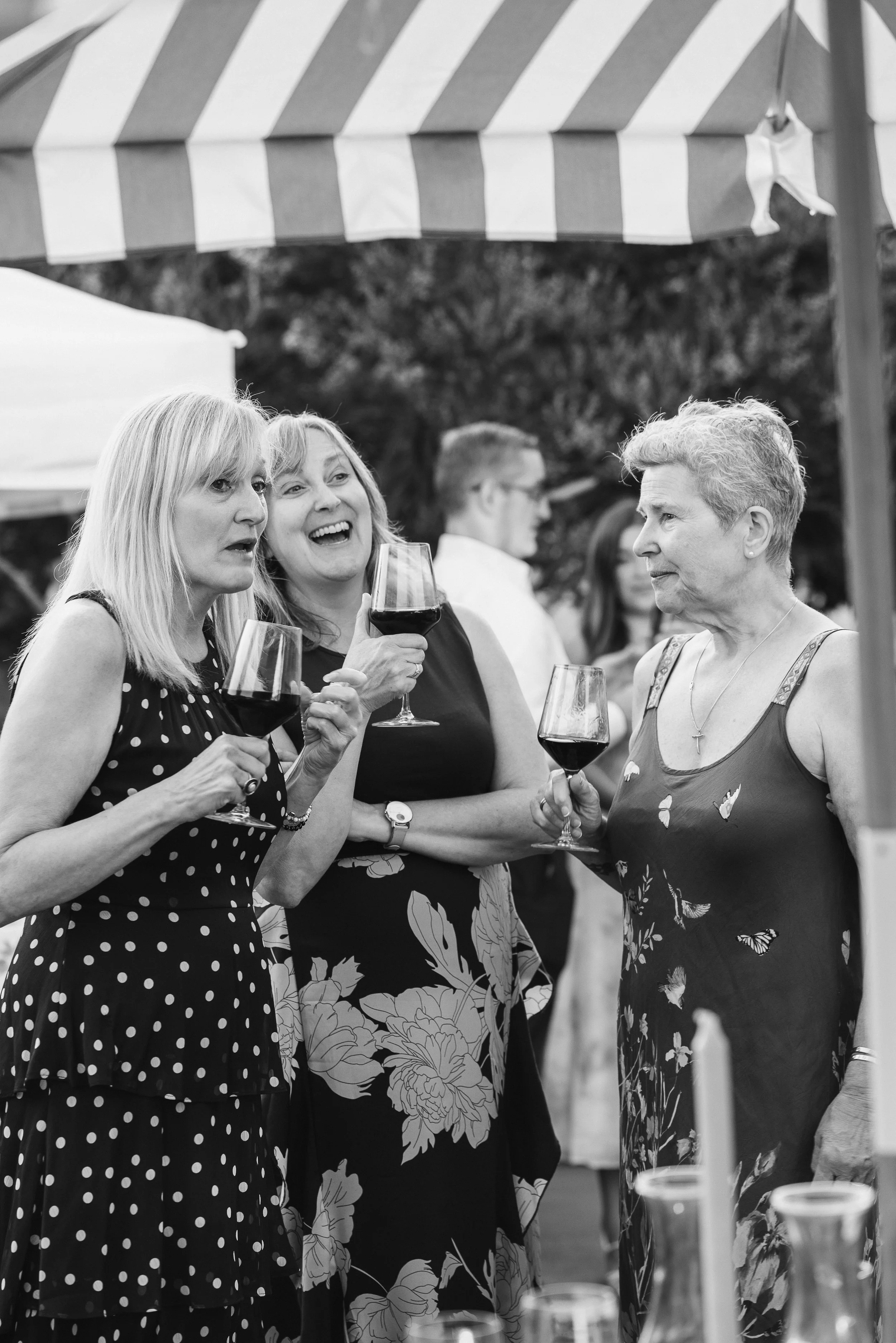Three women holding glasses of red wine, talking and smiling at an outdoor social gathering, with tents in the background.