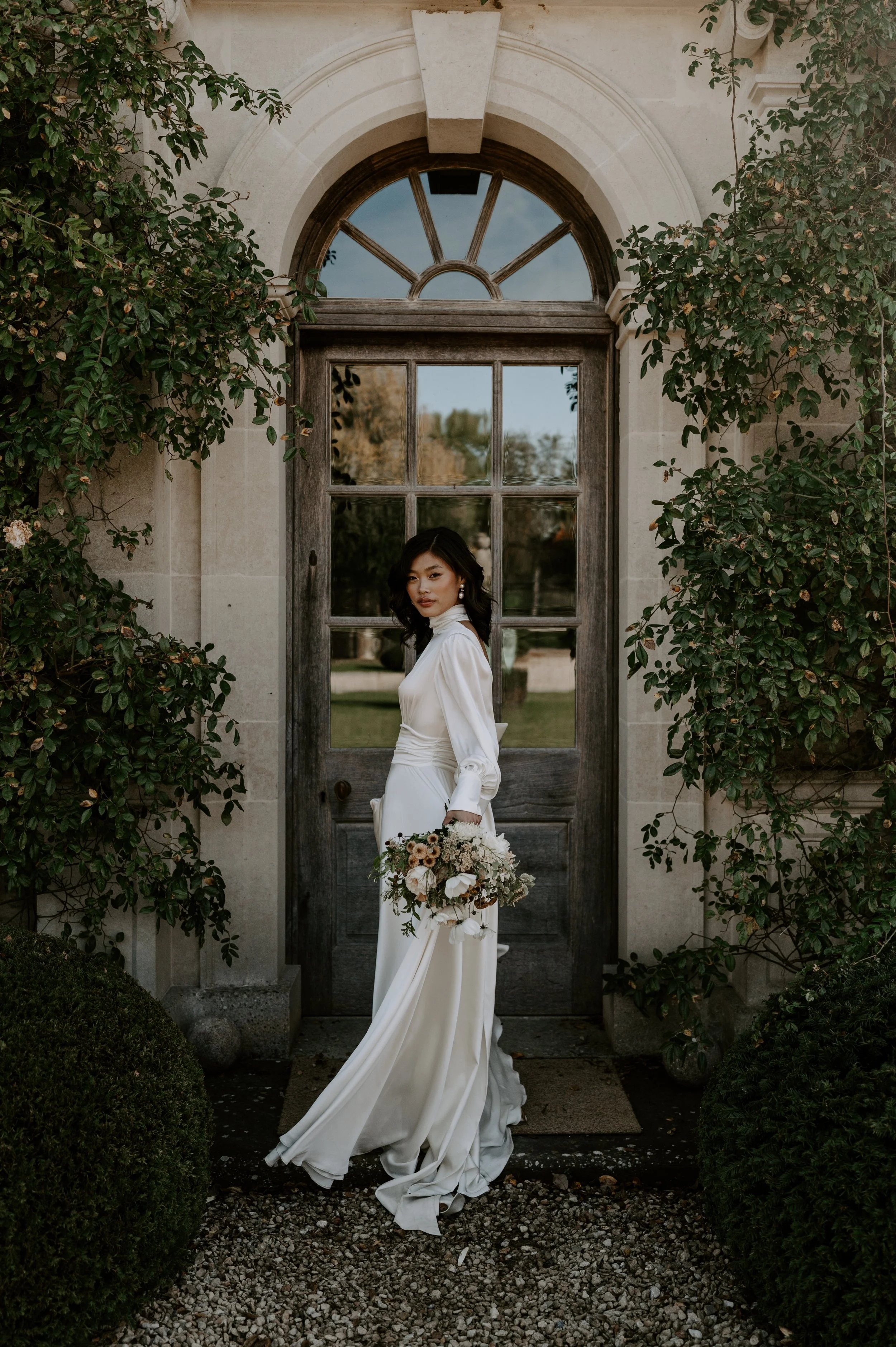 A woman in a white wedding dress holding a bouquet, standing in front of a wooden door surrounded by greenery.
