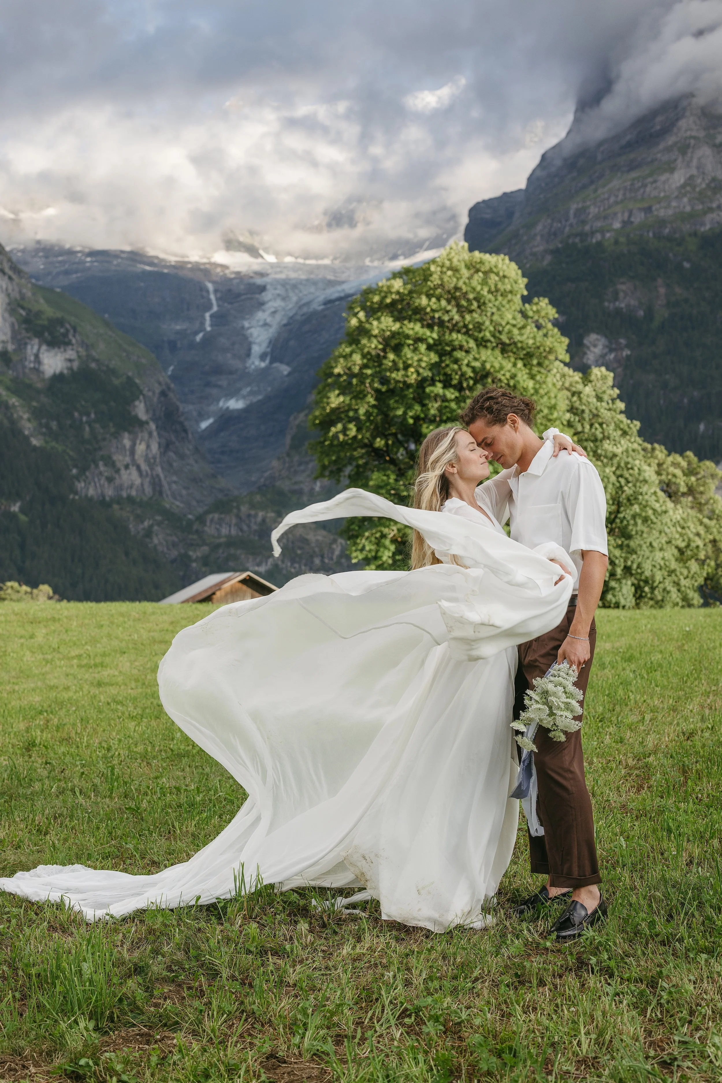 A couple embraces on a grassy field with a mountainous landscape in the background, the woman in a white dress and the man in a white shirt and brown pants, holding a bouquet of flowers.