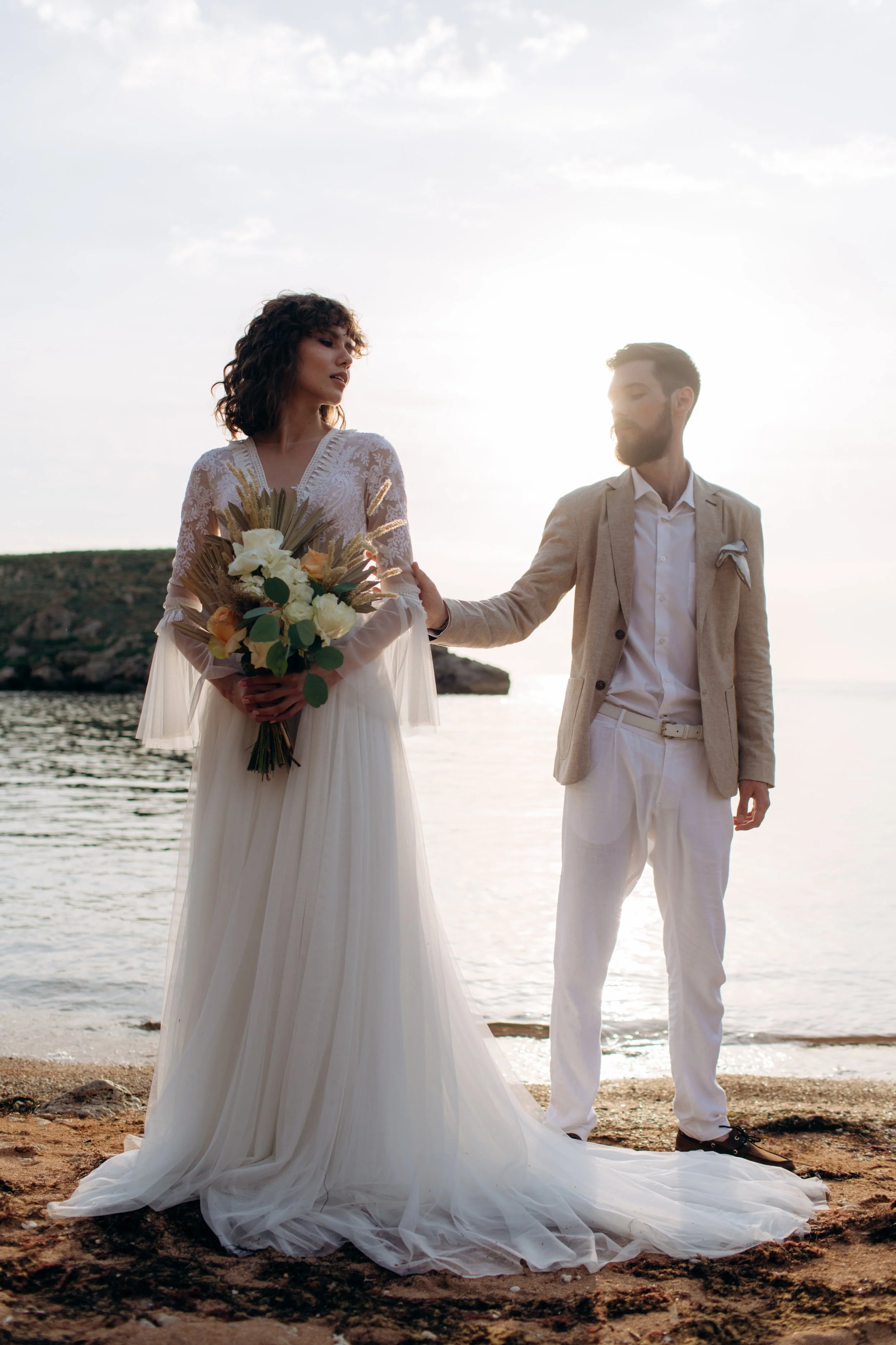 A bride in a white wedding dress holding a bouquet of flowers stands on a beach, with a groom in a tan blazer and white pants touching her arm, during sunset.
