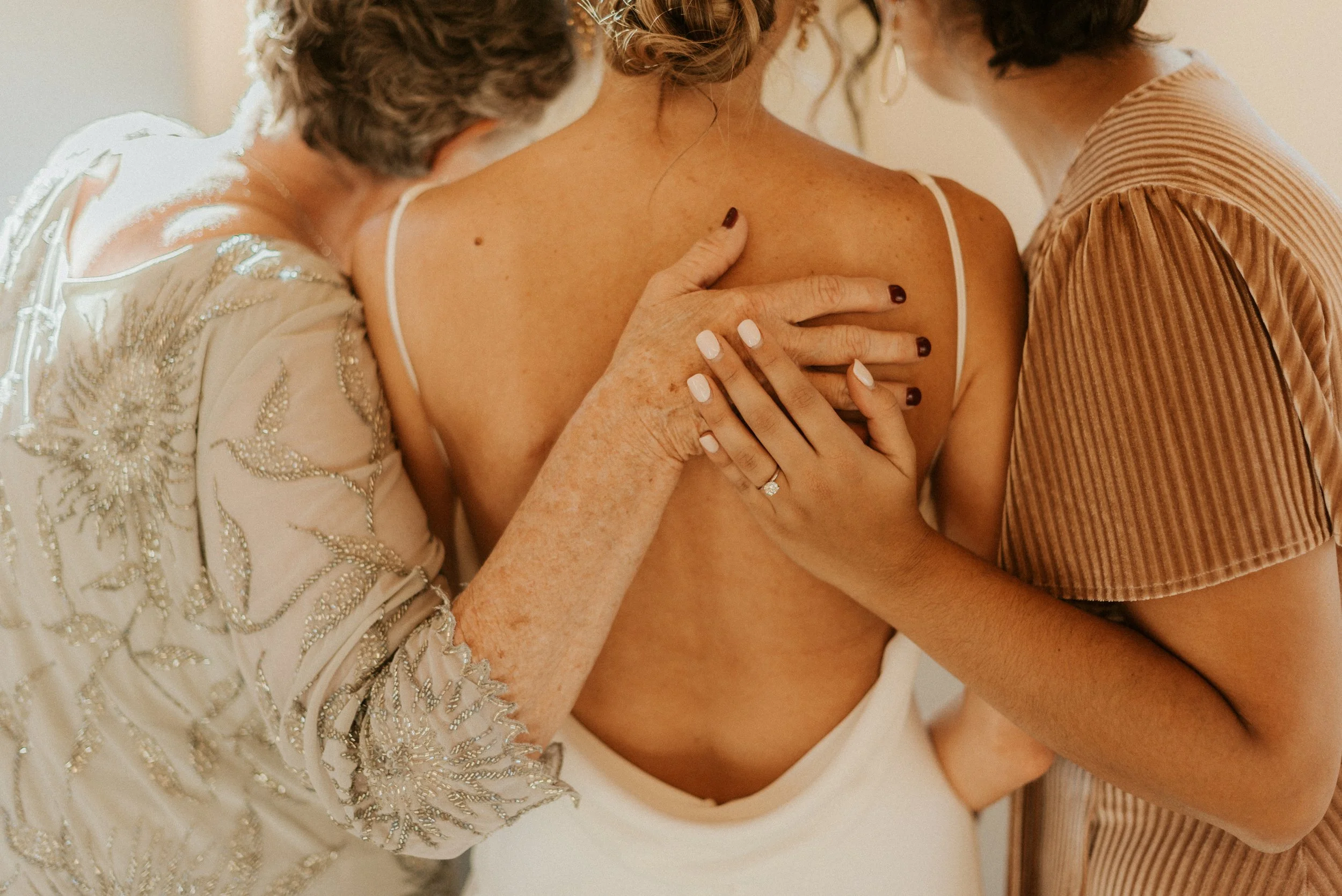Three women embracing, with two women holding and touching the back of a young woman in a white dress, focusing on the intimate and emotional moment.