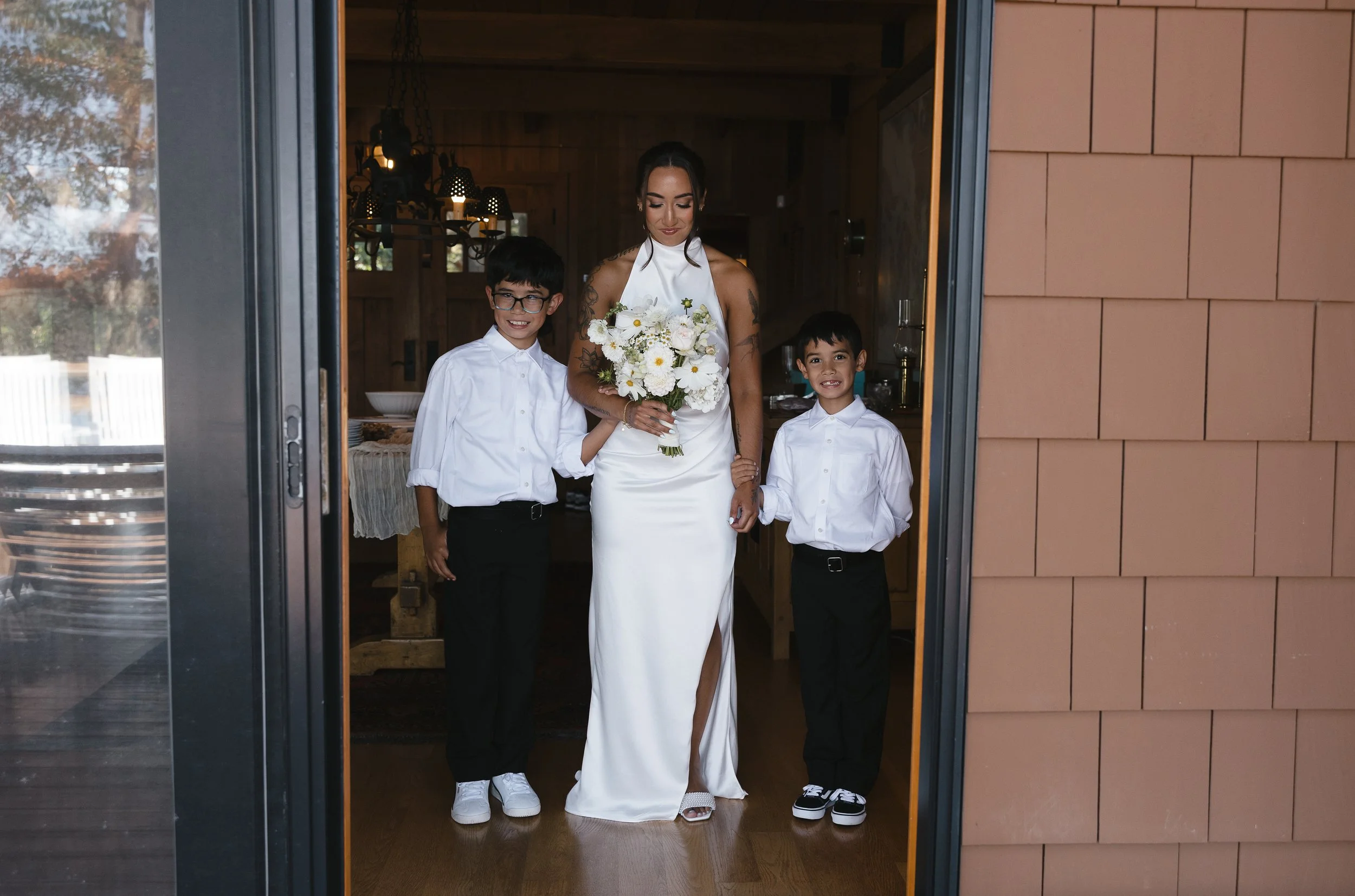 A woman in a white dress holding a bouquet of flowers standing in a doorway with two boys in white shirts and black pants on either side of her, inside a warmly lit room.