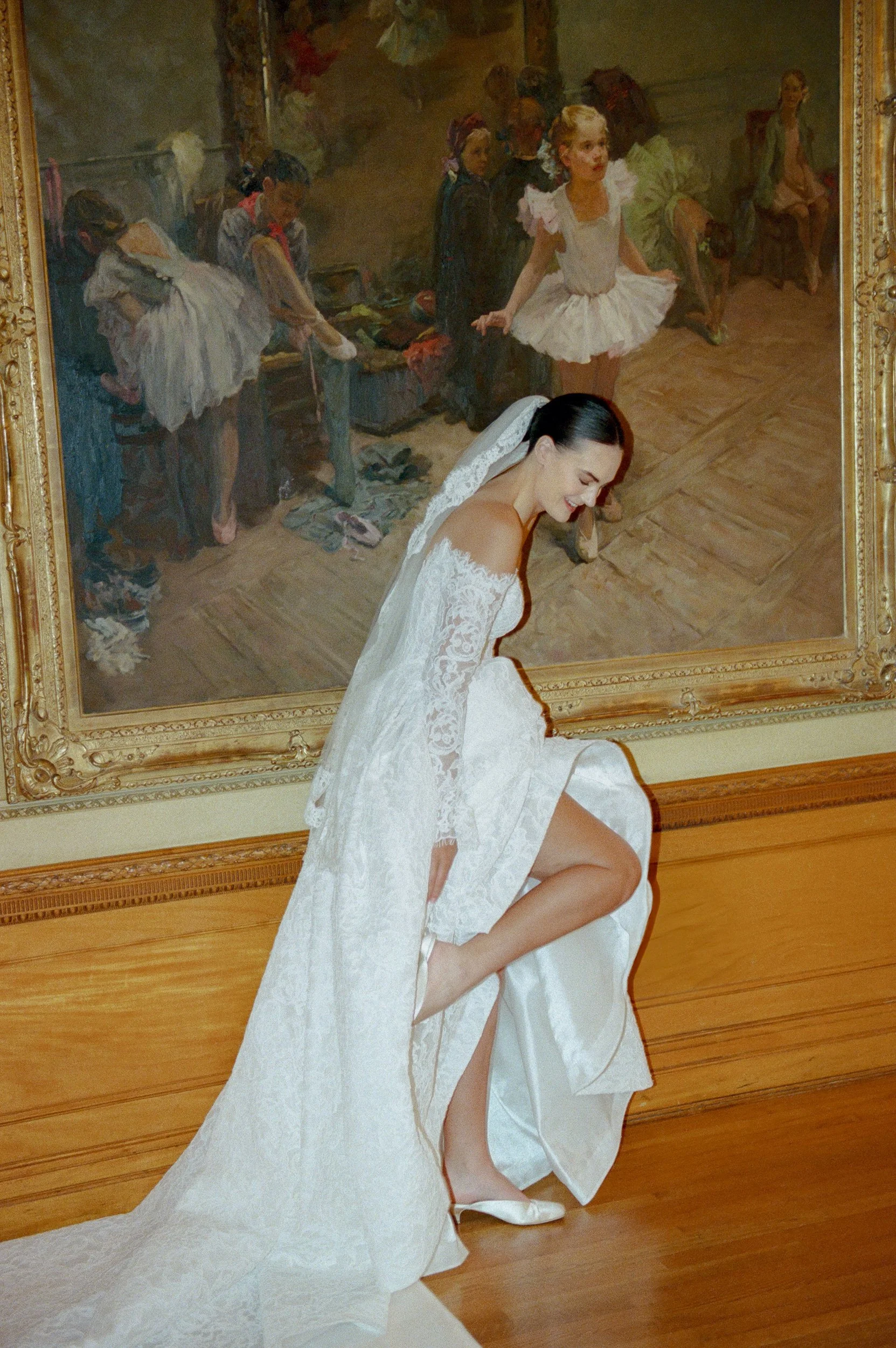 A woman in a white lace wedding dress and veil adjusting her shoe while smiling in front of a large painting of a ballet scene.