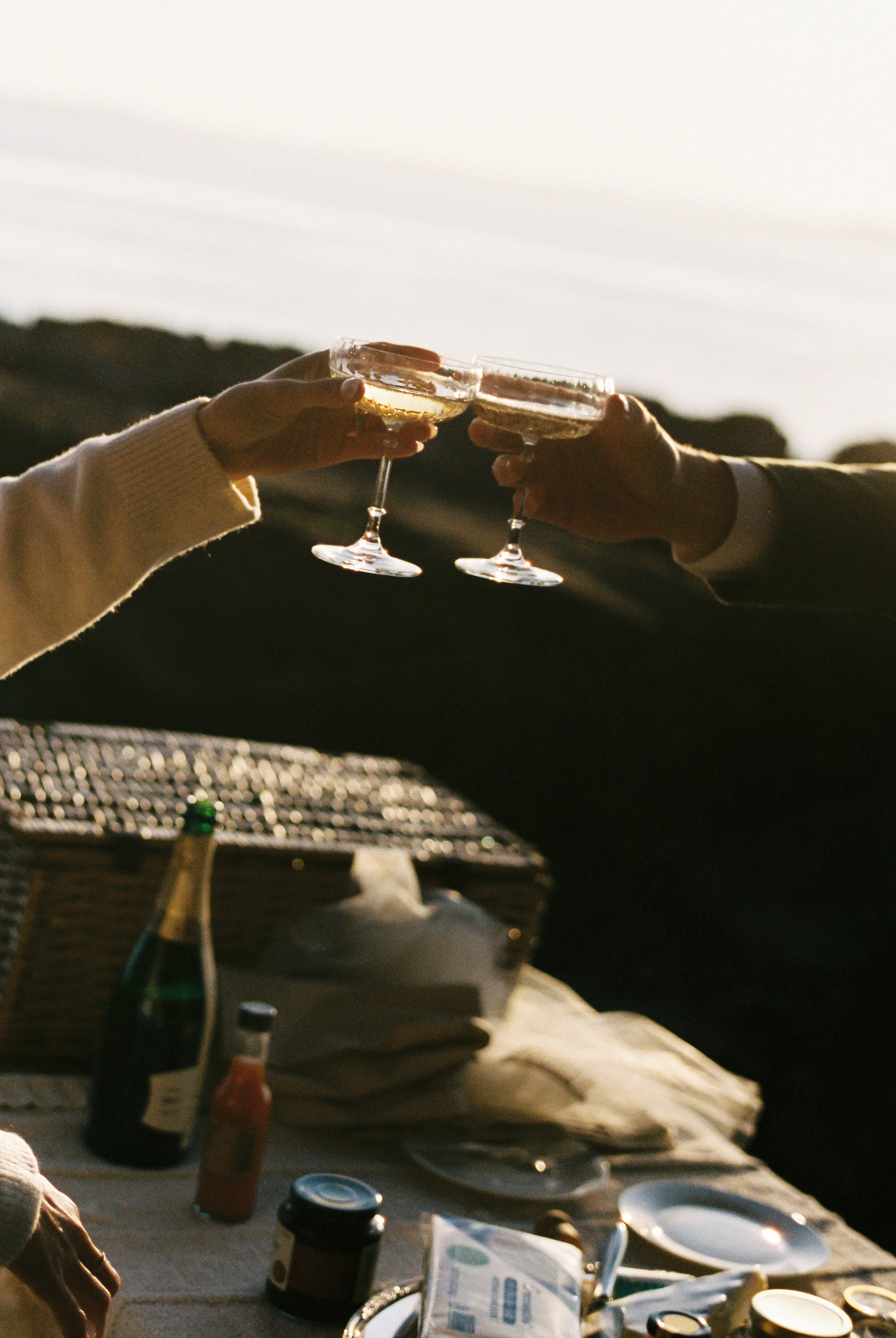 Two people toasting with champagne glasses during a picnic at sunset.