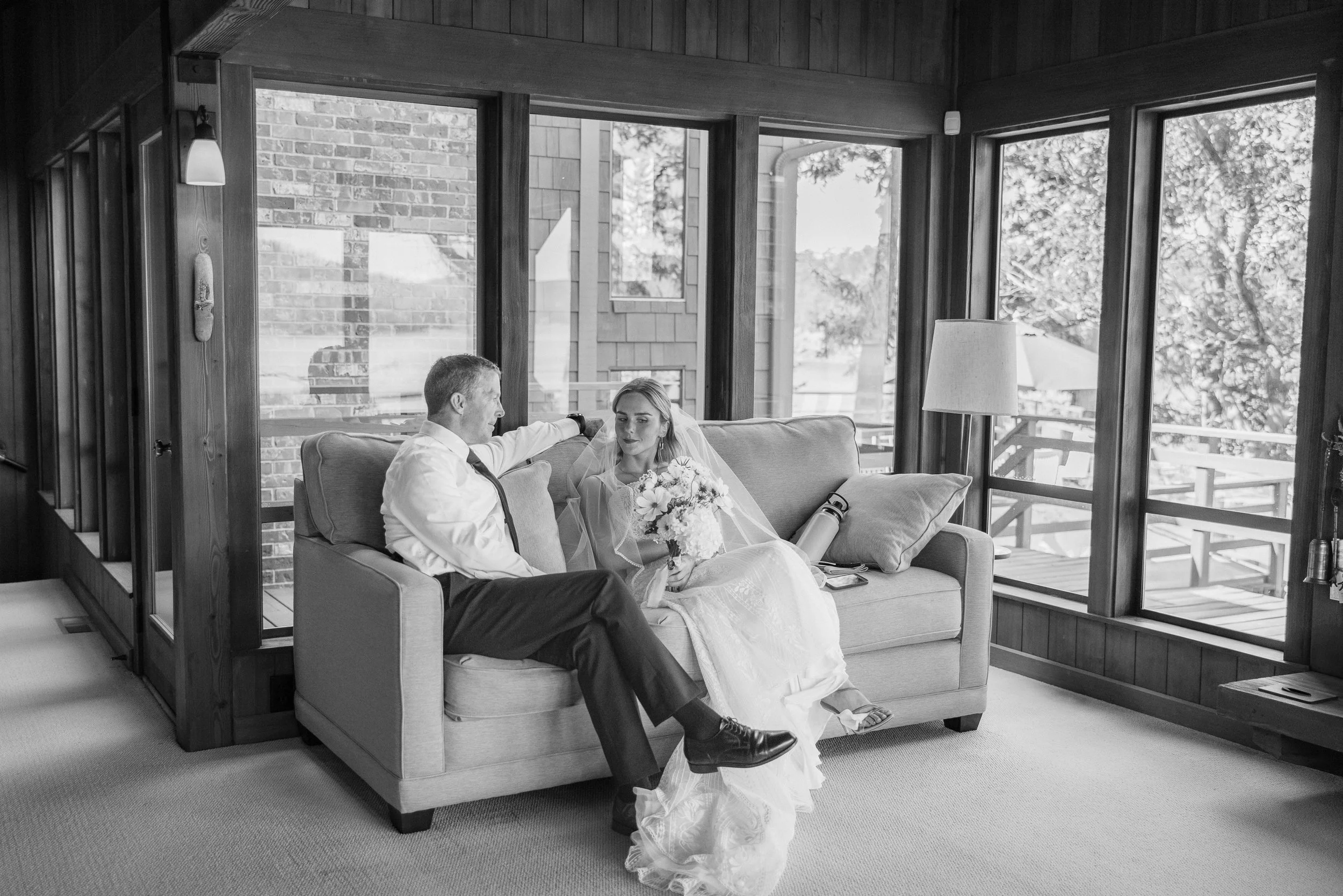 Black and white photo of a bride and groom sitting on a couch inside a room with large windows. The bride is holding a bouquet of flowers and wearing a veil, while the groom is dressed in a shirt and tie, leaning back and talking to her.