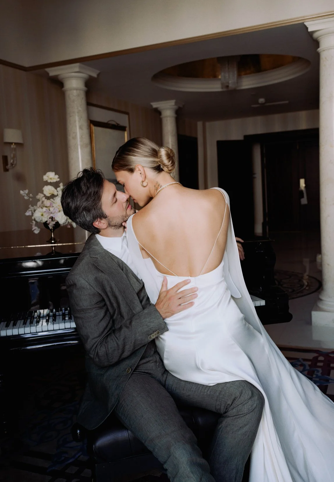 A man and woman in wedding attire sharing an intimate moment in a luxurious room with a grand piano and floral decor.