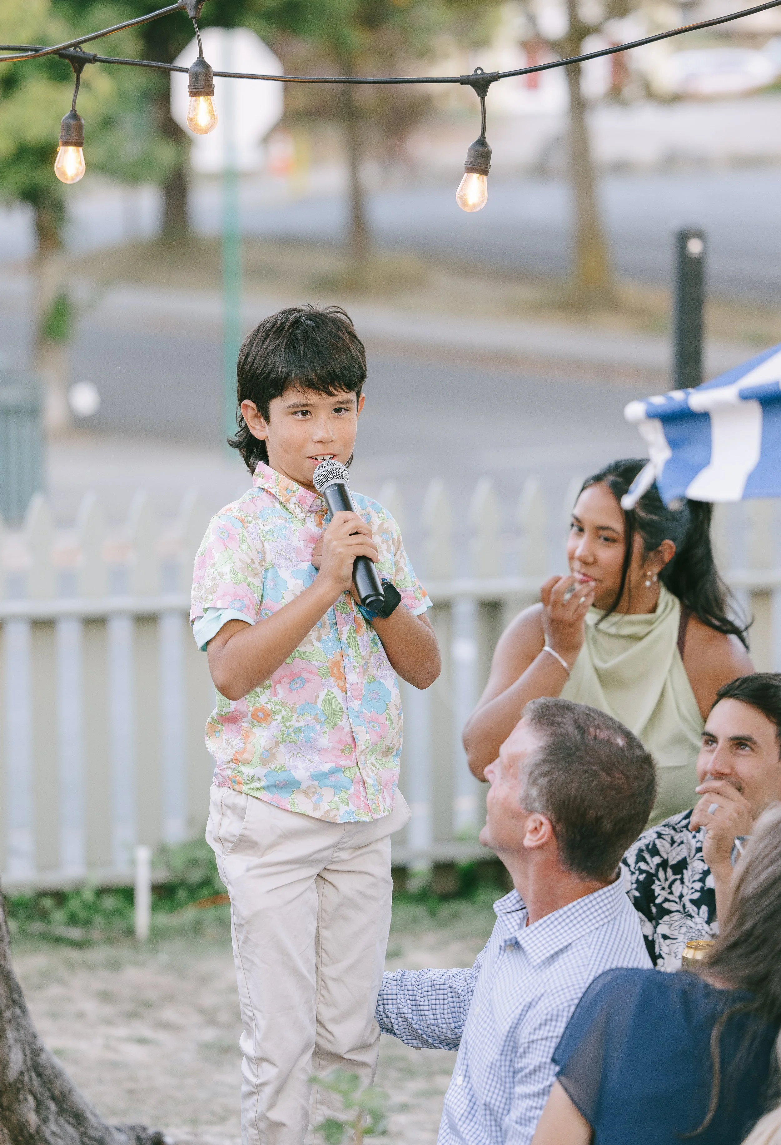 A young boy with dark hair, wearing a floral shirt, stands outdoors holding a microphone. Adults around him are smiling and looking at him, with a woman in a light green dress and two men, one older with short gray hair and one younger with dark hair