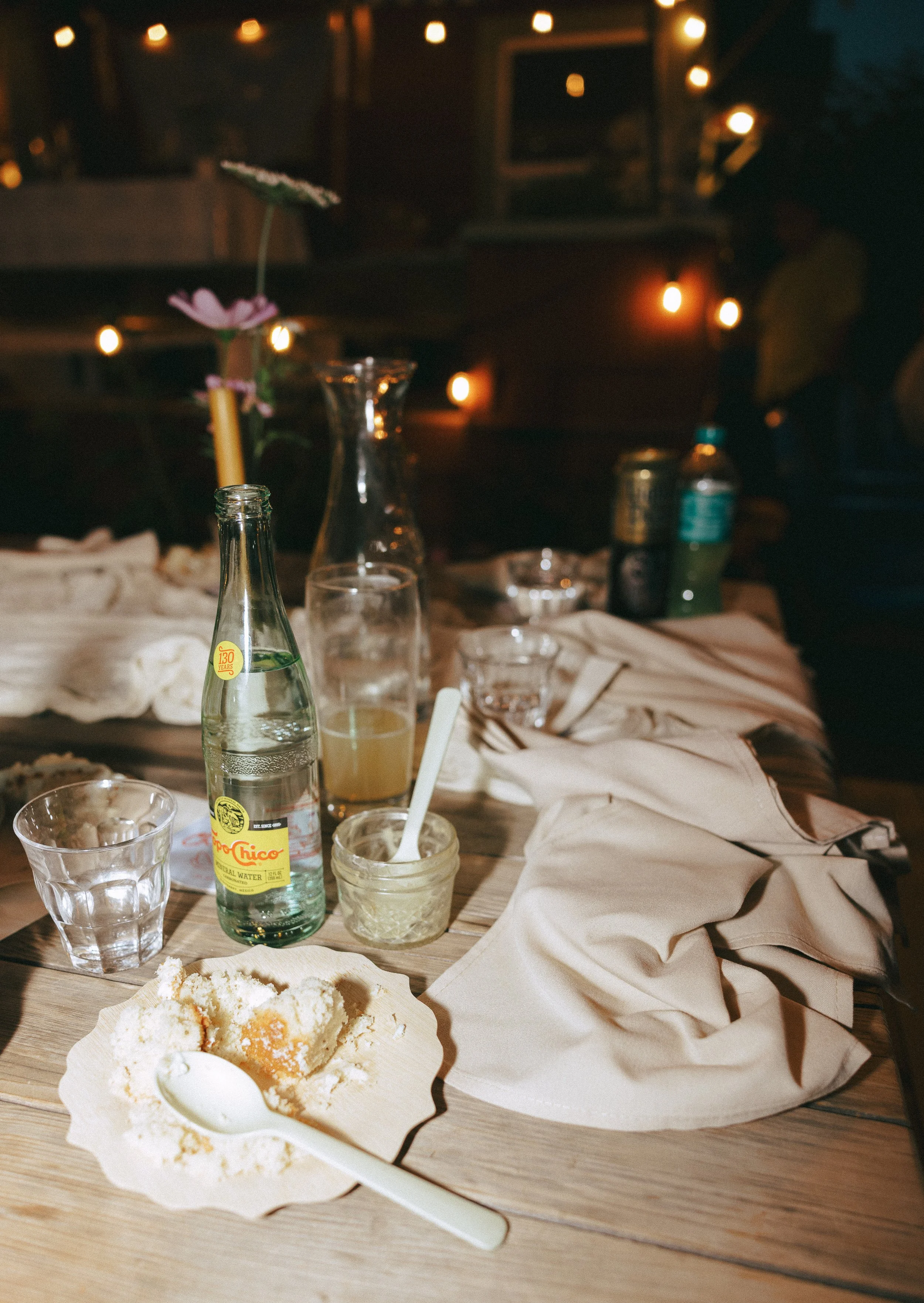 Partially eaten cake on a plate with a spoon, beverage bottles, glasses, and a napkin on a wooden table with blurred background of string lights