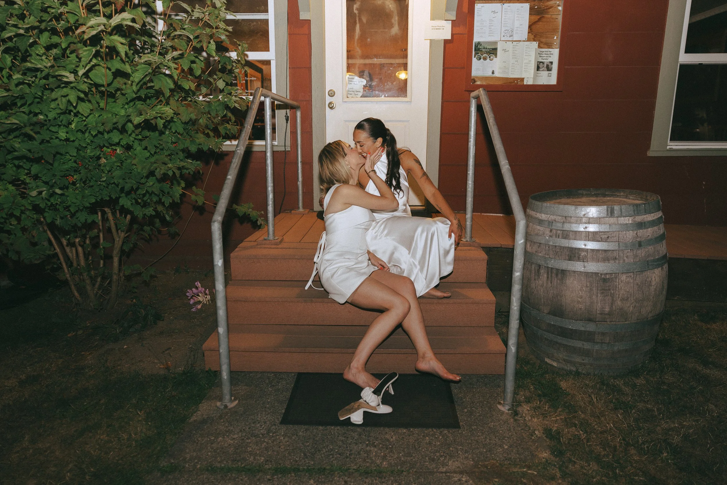 Two women in wedding attire sitting on the steps outside a rustic building, sharing a kiss. One woman is in a white dress, and the other in a white top and shorts. A pair of high-heeled shoes is on the ground nearby, and a large wooden barrel is on t