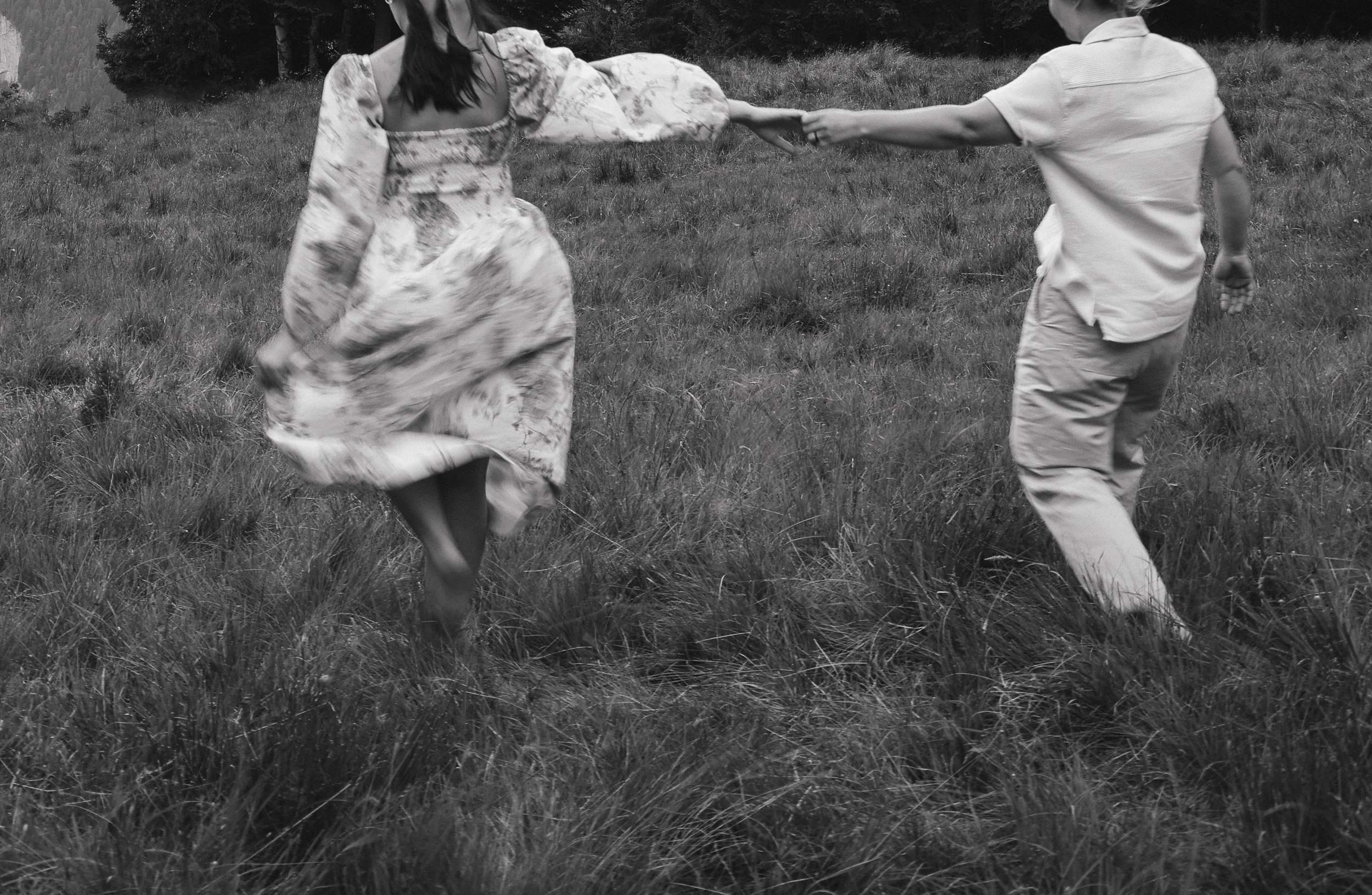 A black and white photo of a man and woman holding hands and running through a grassy field, with trees in the background.