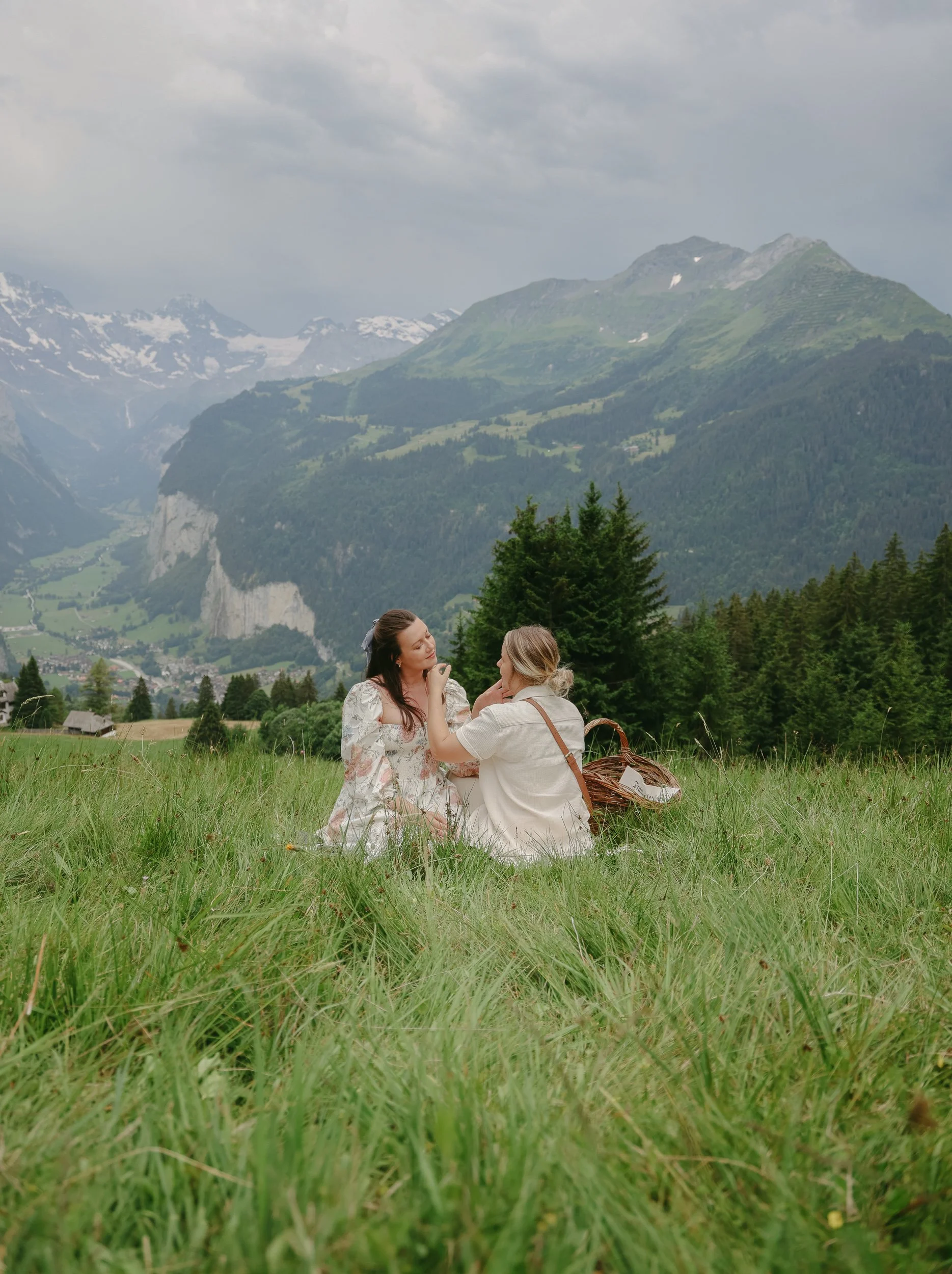 Two women sitting on a grassy field with mountains in the background, one woman easing the other's makeup or face, with a picnic basket nearby.
