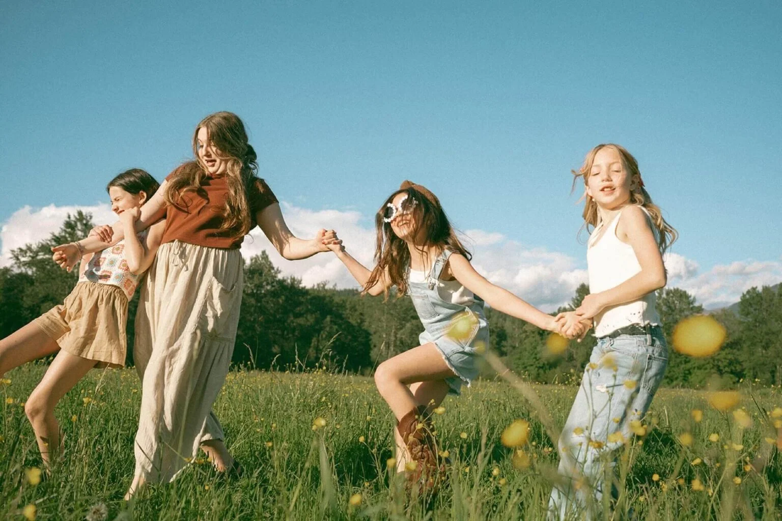 Four girls holding hands and playing in a grassy field with wildflowers, under a blue sky with scattered clouds.