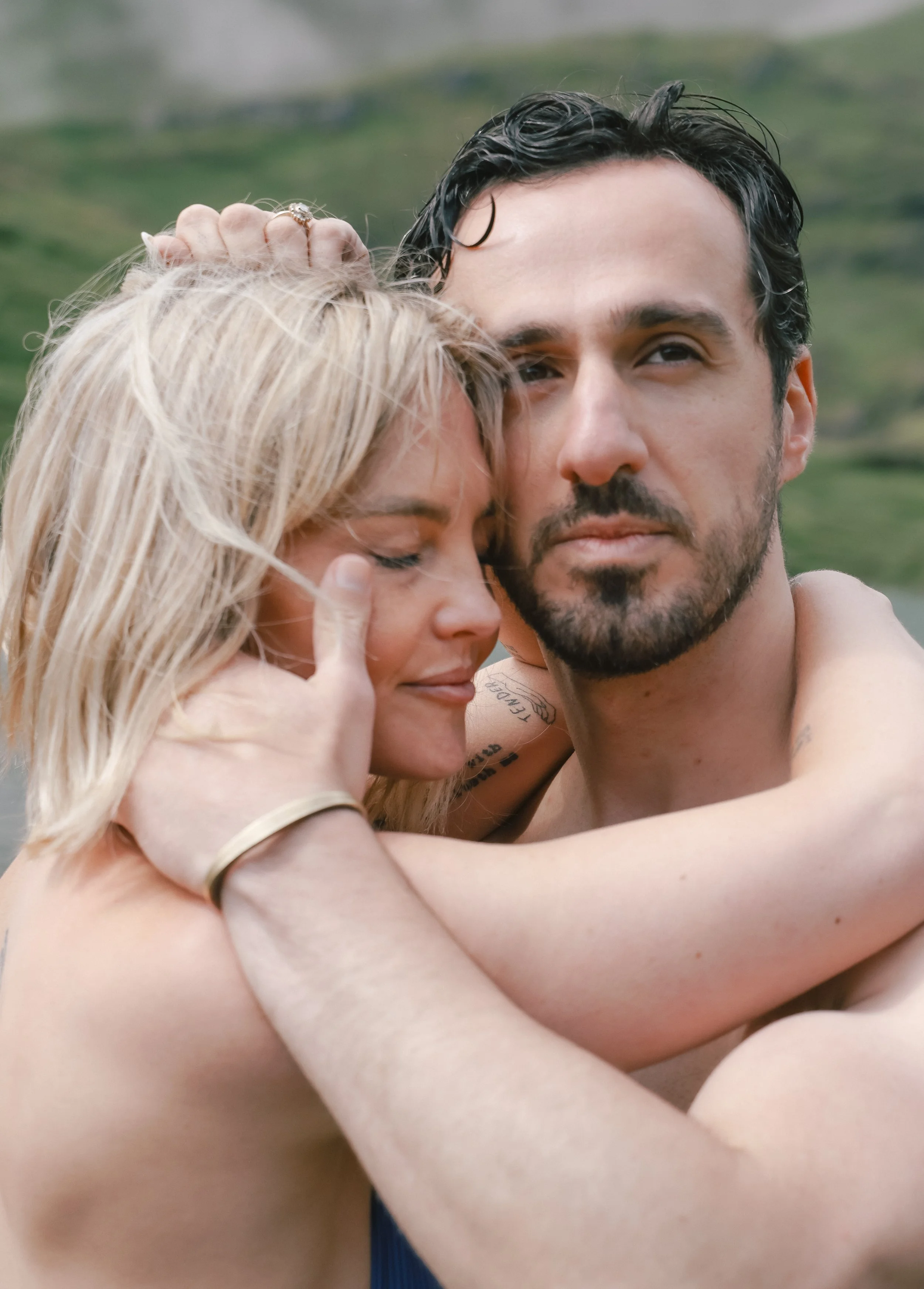 A close-up of a couple hugging outdoors, with hills and greenery in the background. The woman has blonde hair, and the man has dark hair and a beard.