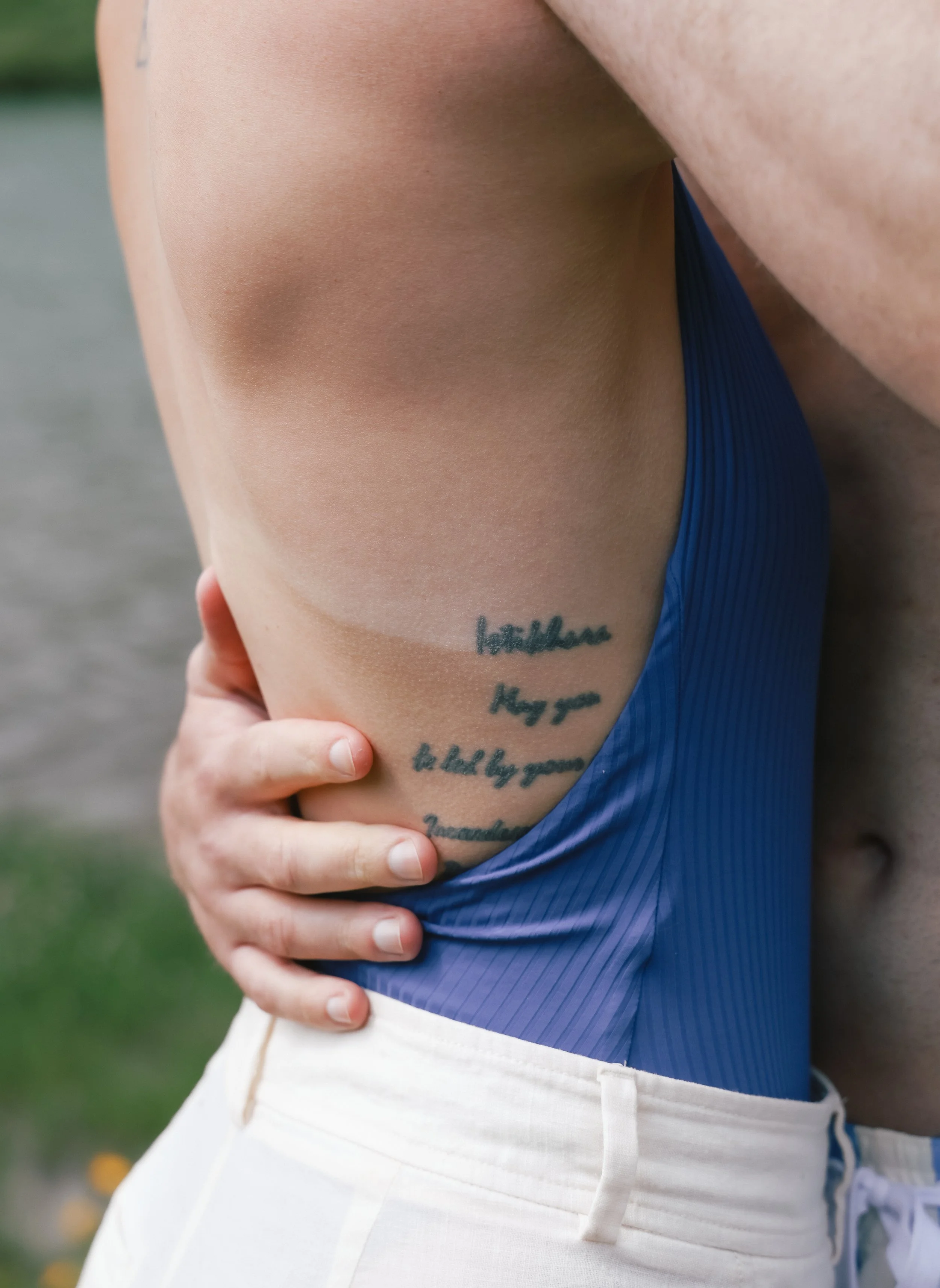 Close-up of a person's side torso, wearing a blue swimsuit, showing a tattoo with handwritten-style text.