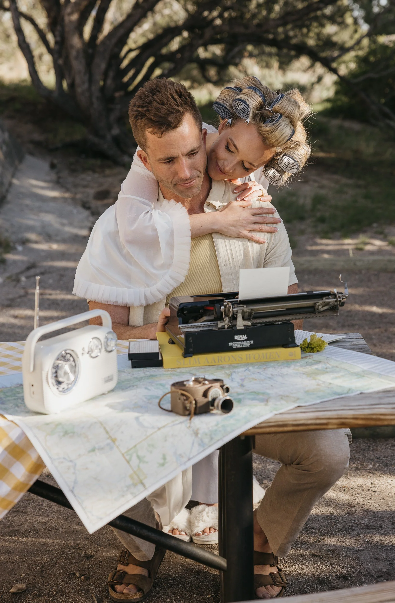 A man and woman sitting at a picnic table outdoors, with the woman hugging the man from behind. The woman has hair rollers, and the man is typing on a typewriter. On the table, there is a radio, a camera, and a map.
