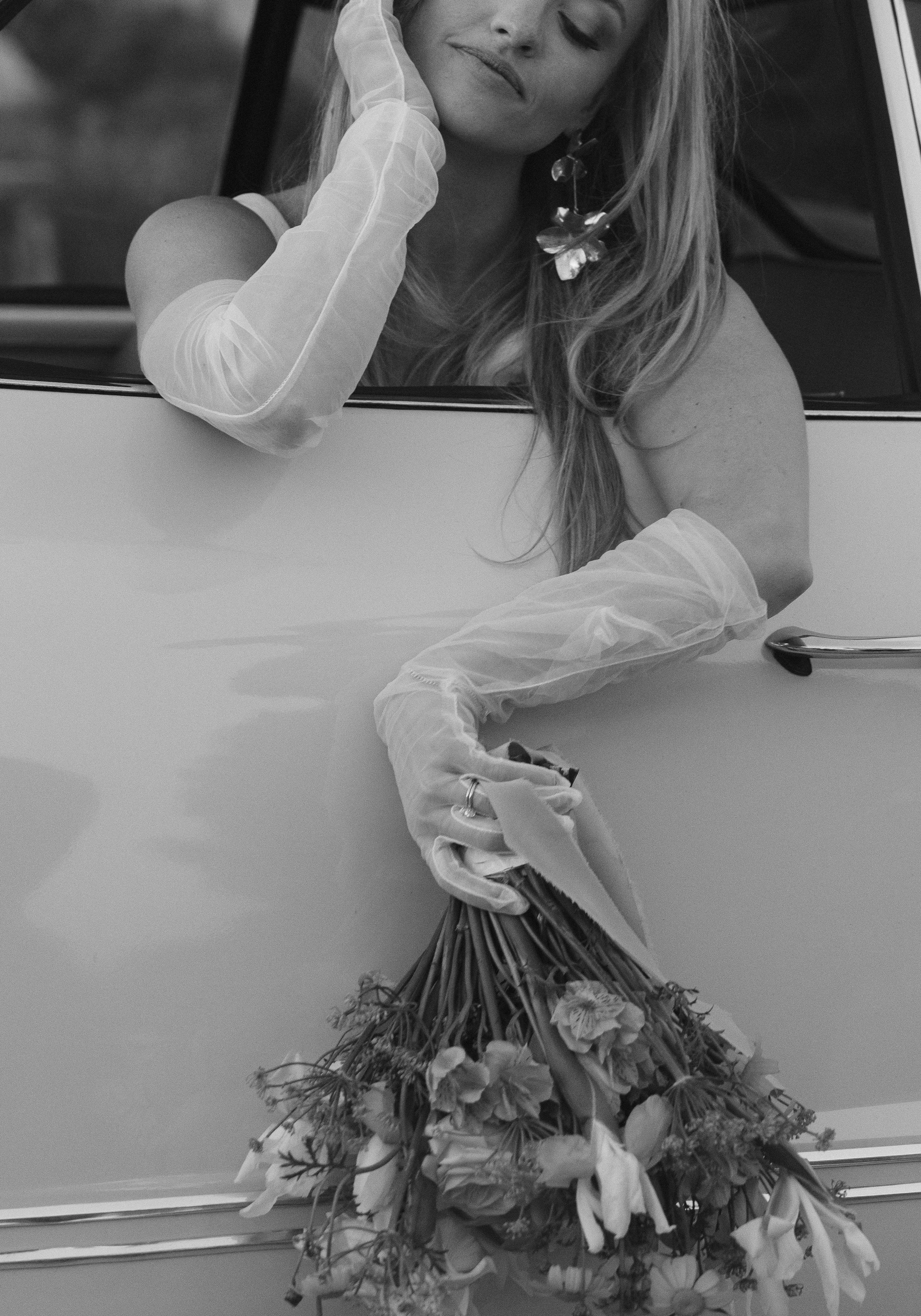 A woman with long hair and earrings sitting in a vintage car, holding a bouquet of flowers.