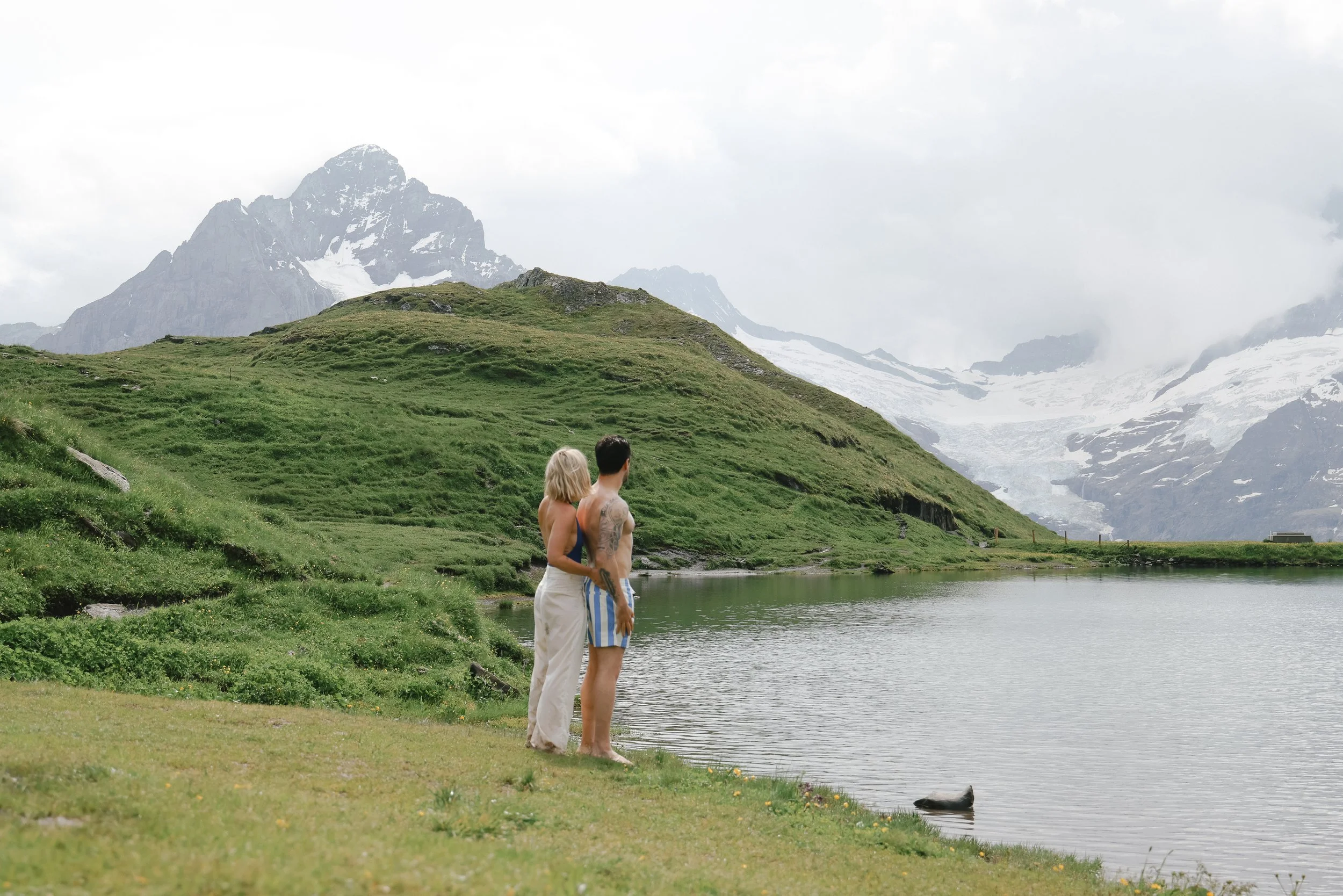 A man and woman standing by a lake in a mountainous area with snow-capped peaks and green hills.