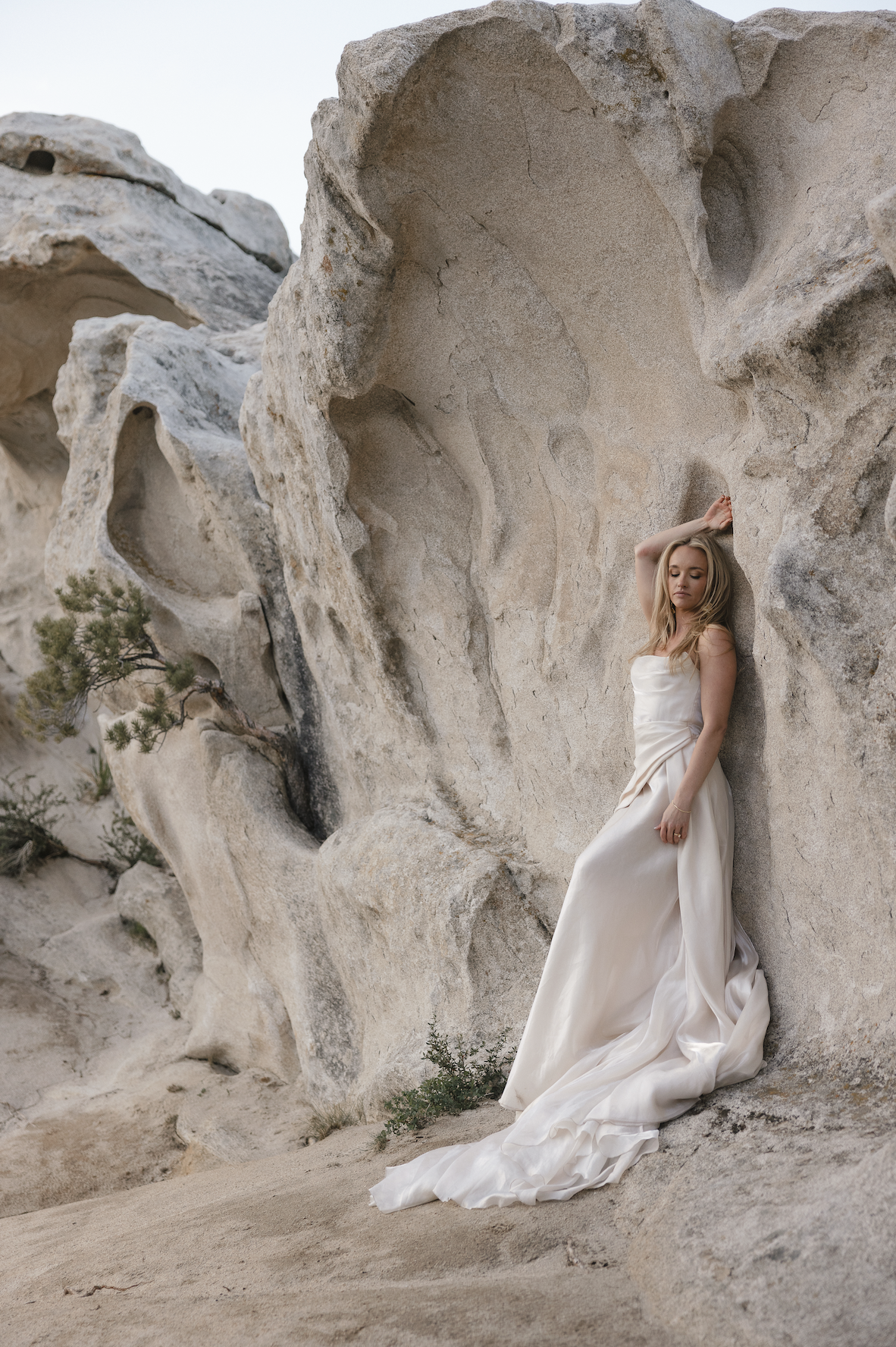 A woman in a white wedding dress leaning against a large, textured rock formation in a desert landscape with small green plants.