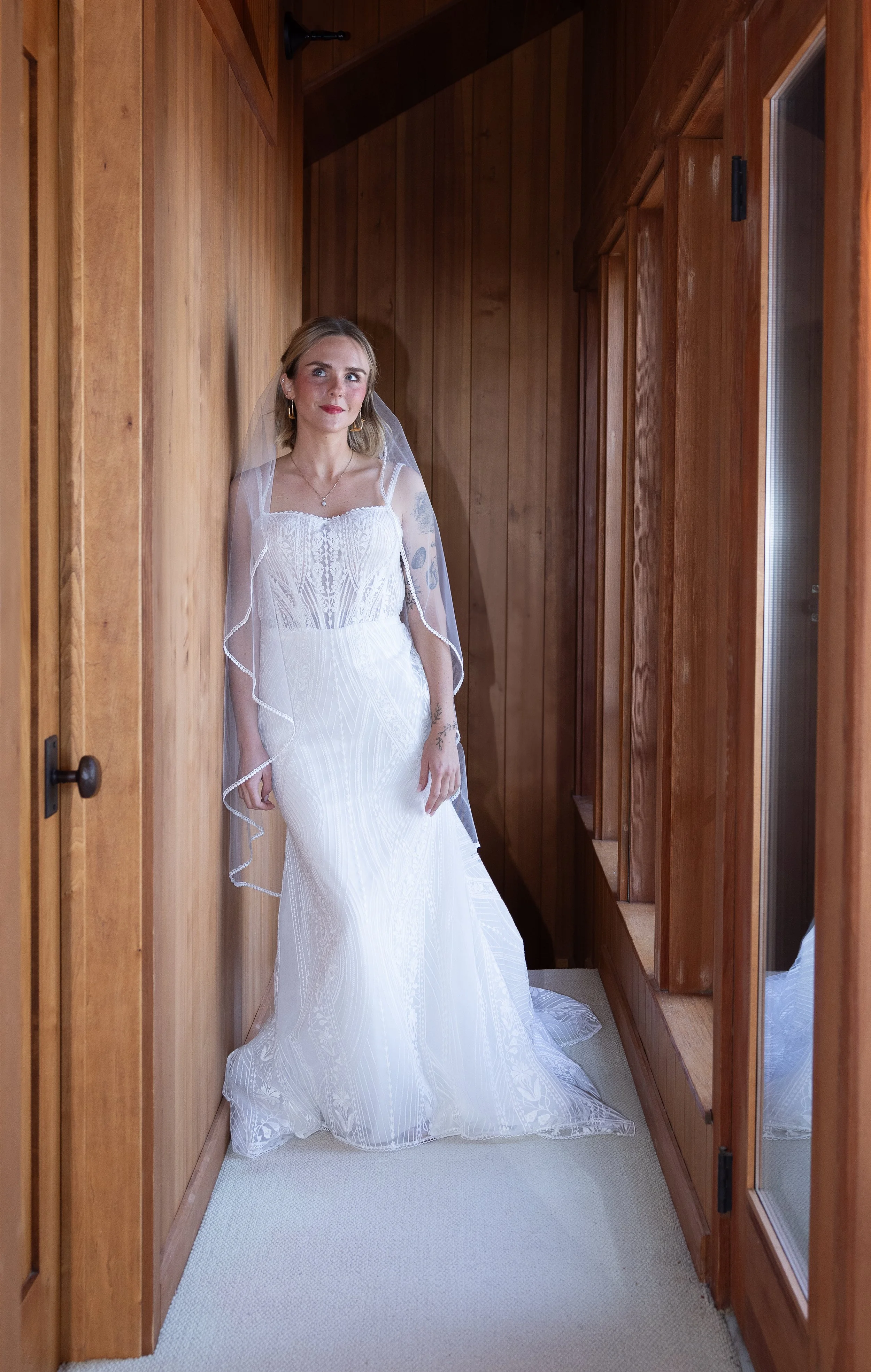 A bride in a white wedding dress with lace details, standing in a wooden-paneled hallway next to a window and looking upward.