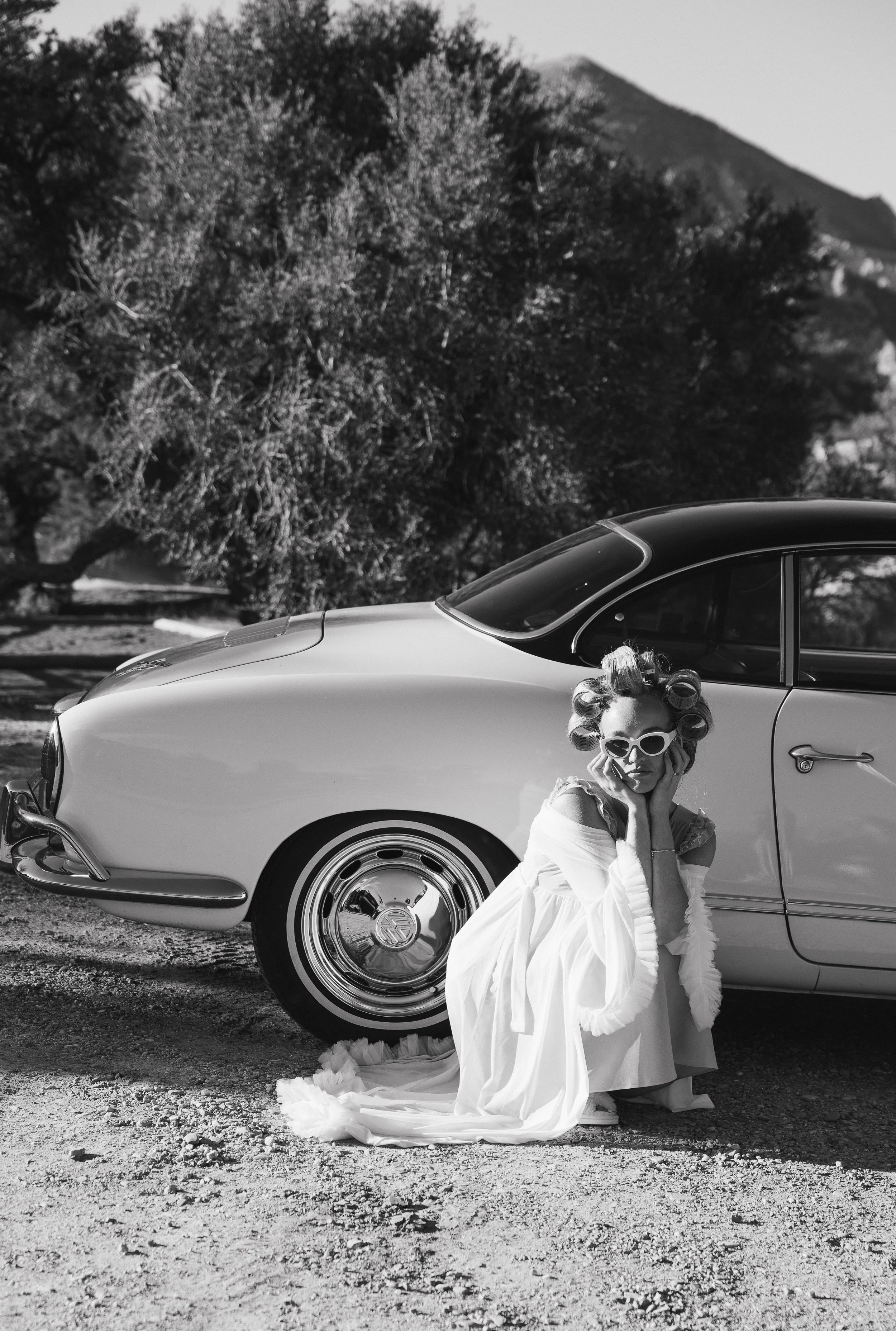 A woman dressed in vintage style with curlers and sunglasses, sitting on the ground beside a classic car with a scenic mountain and trees in the background.