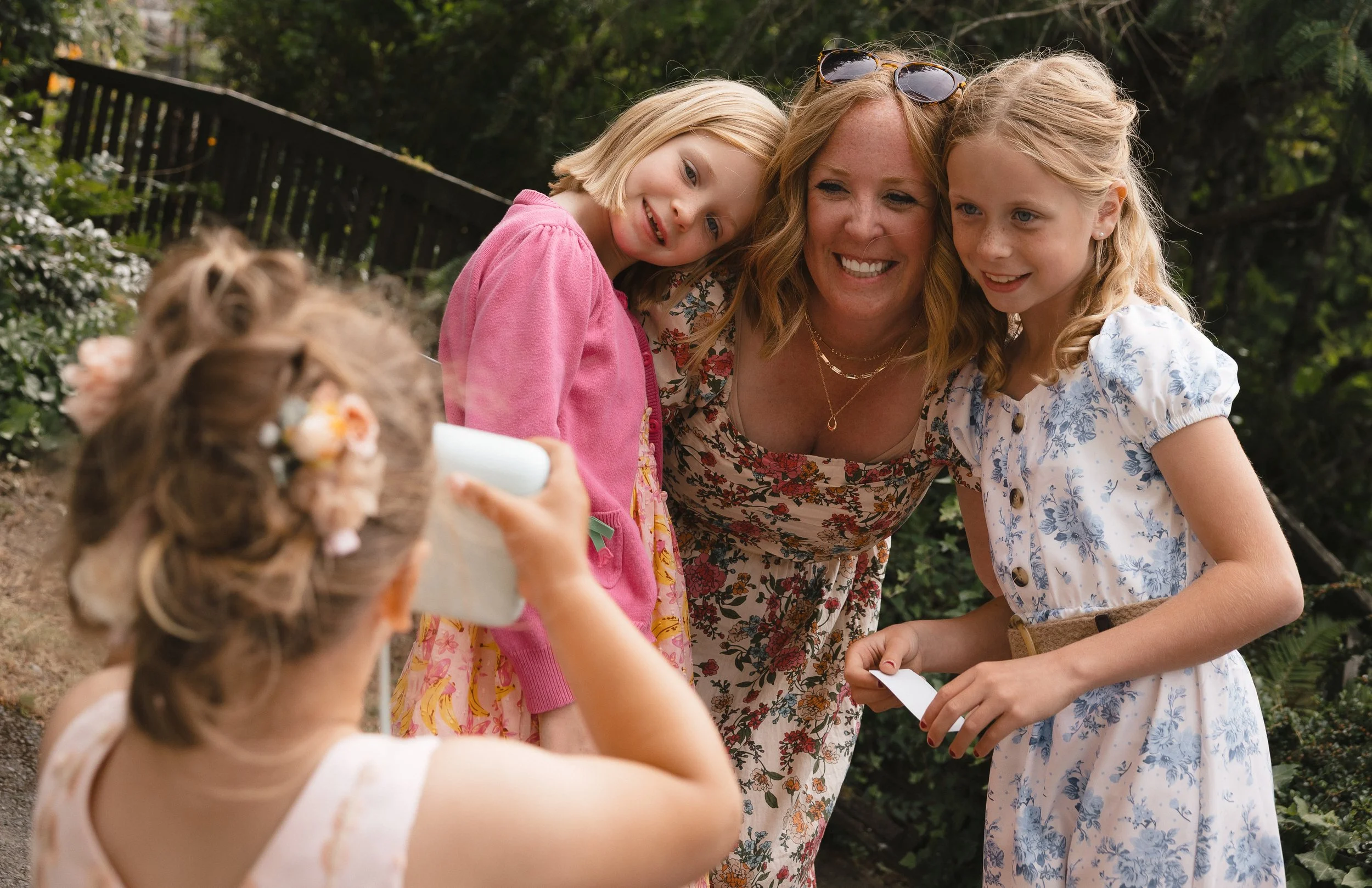 A woman with blonde hair and sunglasses on her head is taking a photo of three young girls with a smartphone. The girls are smiling and posing outdoors in a garden with trees and a wooden fence in the background.