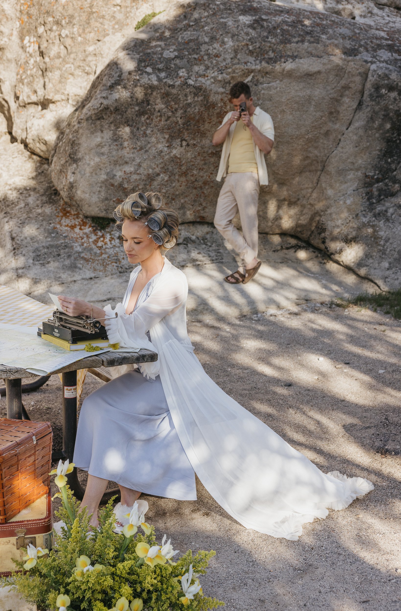 A woman in a white dress with curlers in her hair is sitting at an outdoor table, typing on an old-fashioned typewriter, with a basket and flowers nearby. A man in casual clothing and sandals is taking a photo of her against a rocky outdoor setting.