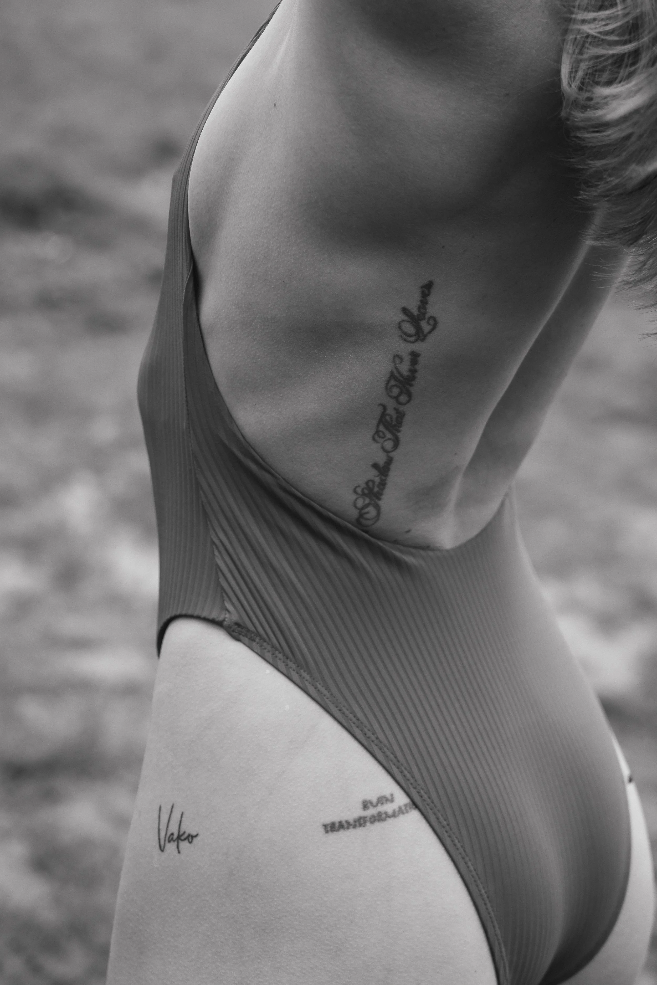 Close-up of a woman's side wearing a swimsuit with tattoos on her ribcage and hip, in black and white.