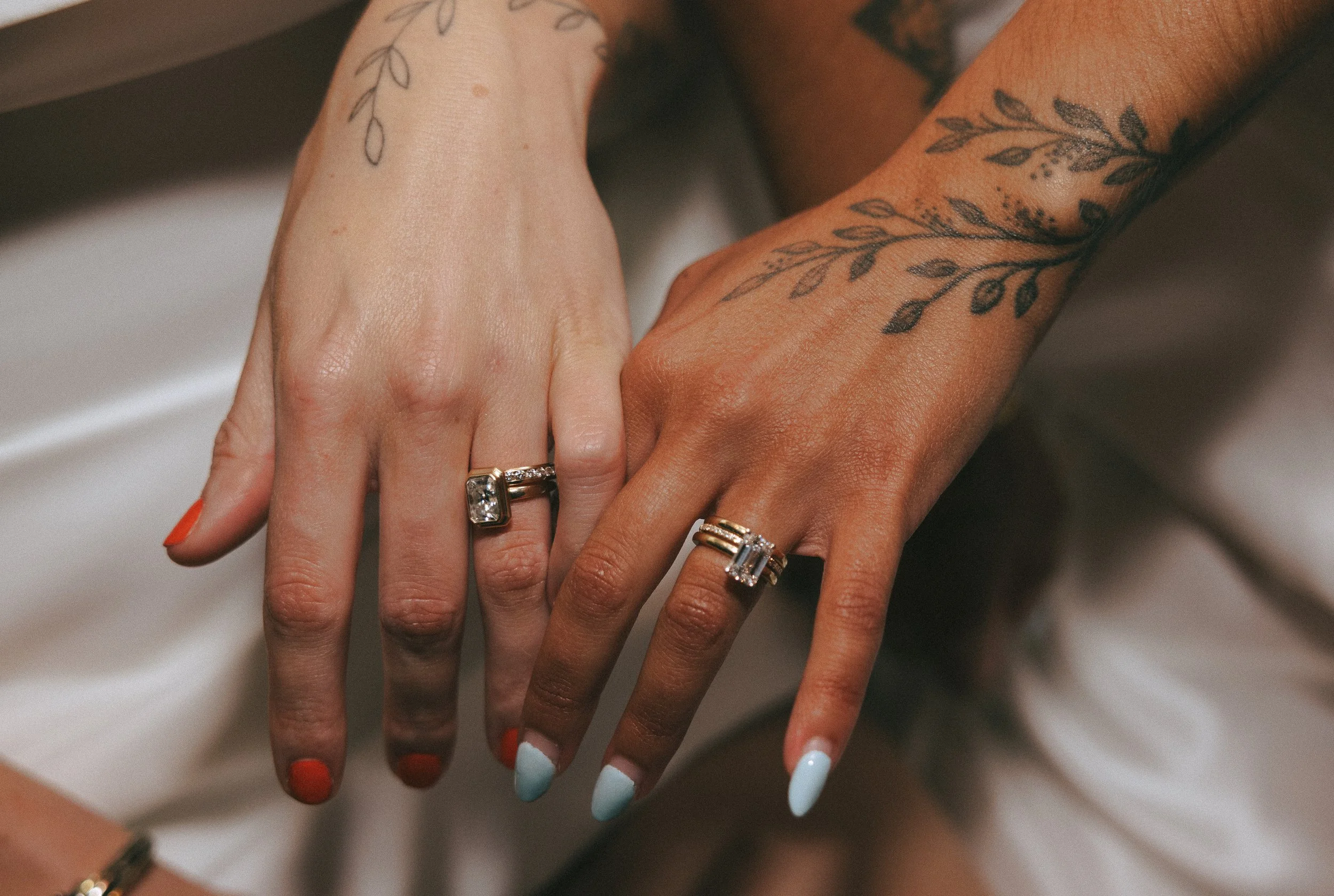 Two hands with wedding rings and floral tattoos, one with red nail polish, and the other with light blue nail polish.