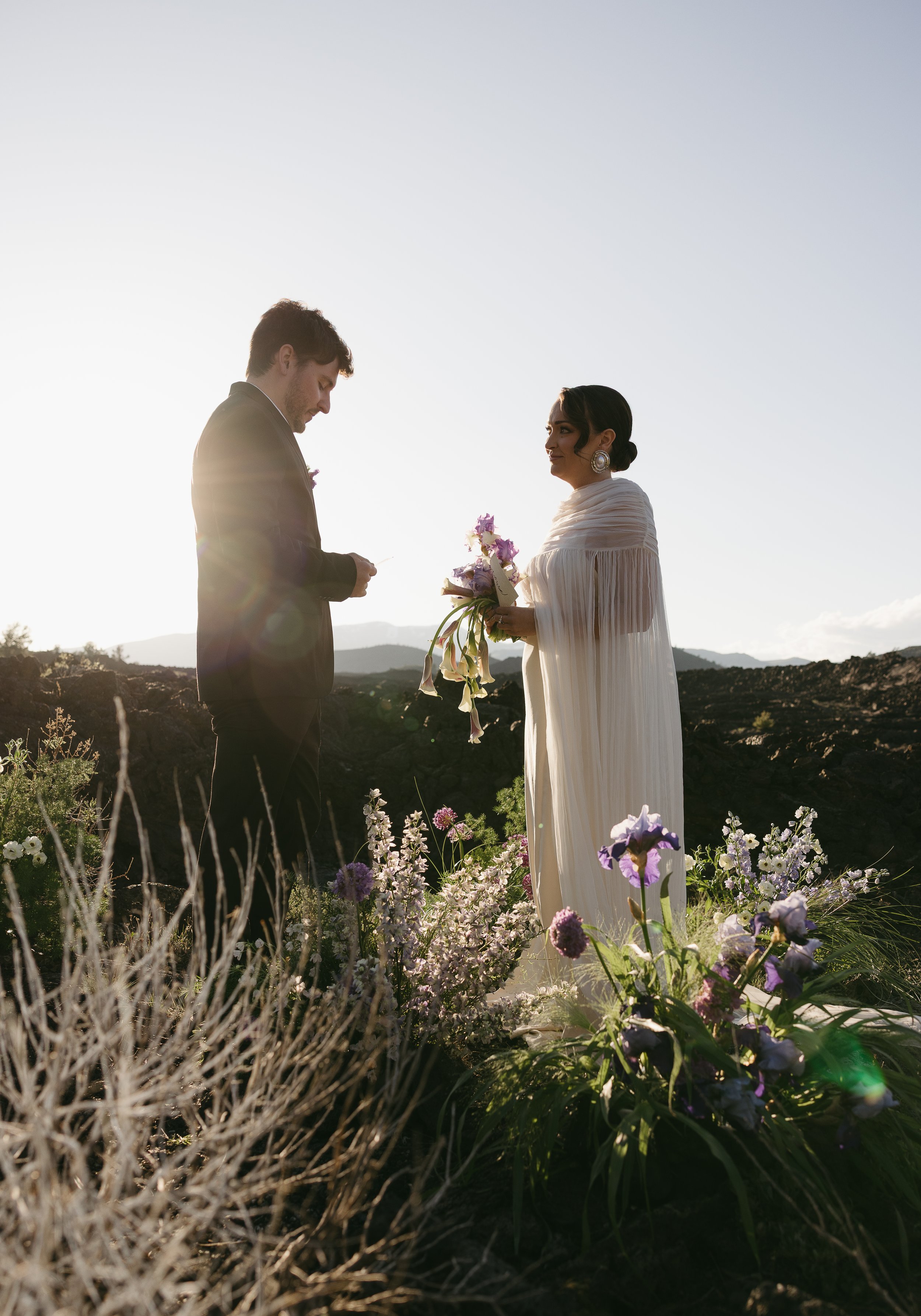A couple during a wedding ceremony outdoors at sunset, with the woman holding a bouquet of purple and white flowers, both in formal attire, surrounded by flowers and plants, mountains in the background.