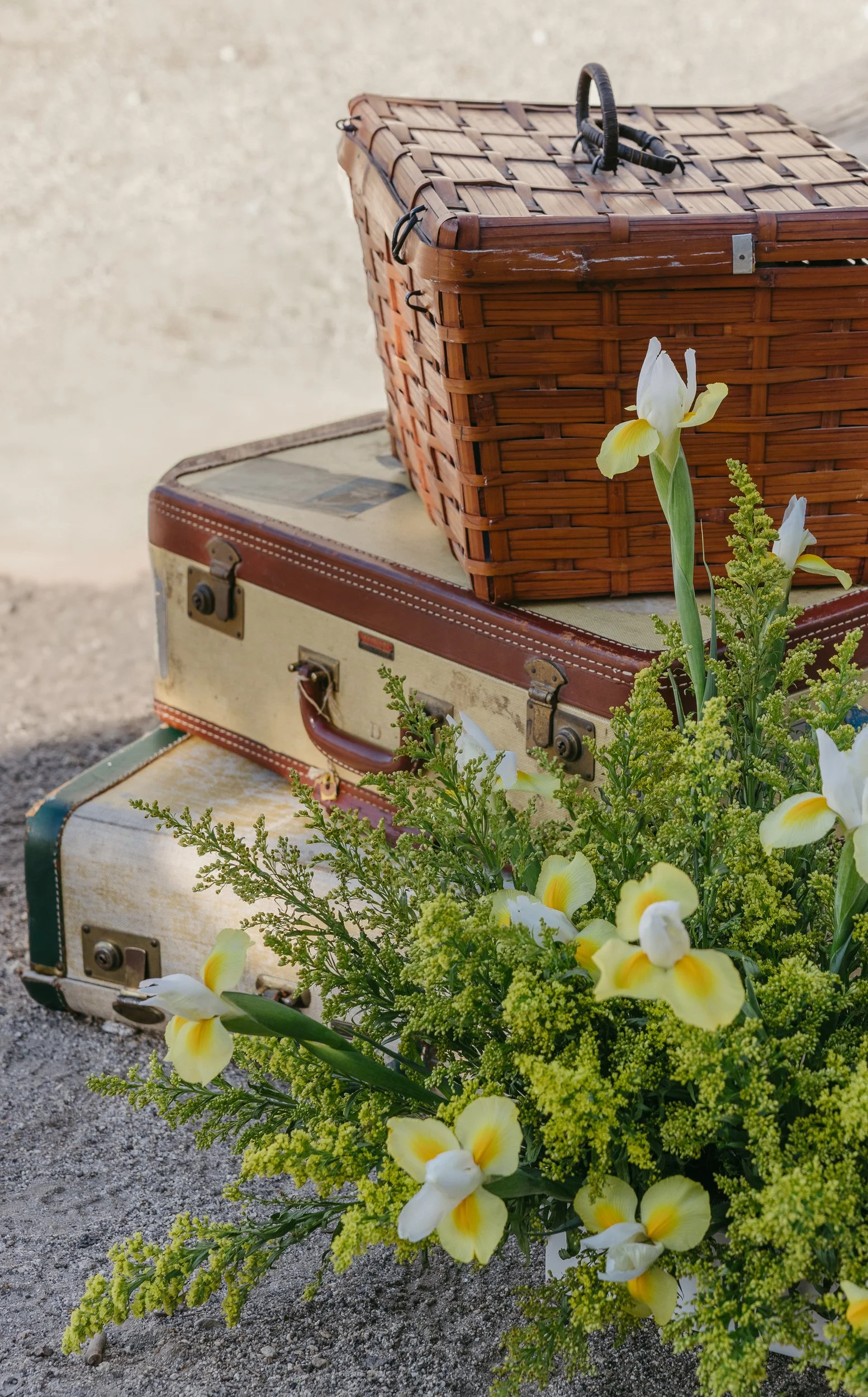 Stack of vintage suitcases with a wicker picnic basket on top, next to a bouquet of yellow and white flowers.