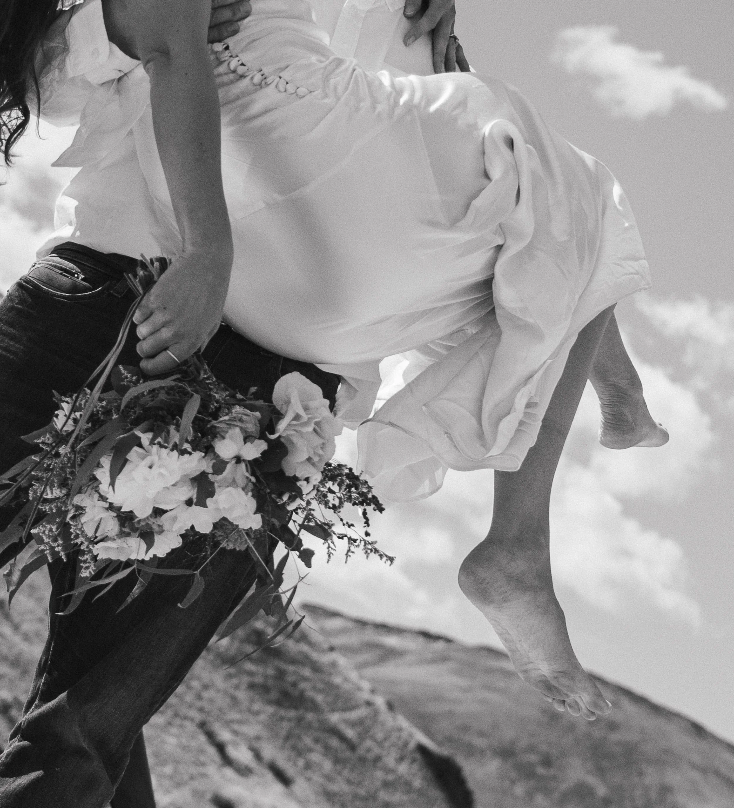 A person holding a woman in a wedding dress, with the woman’s feet lifted off the ground, holding a bouquet of flowers, outdoors under cloudy sky.