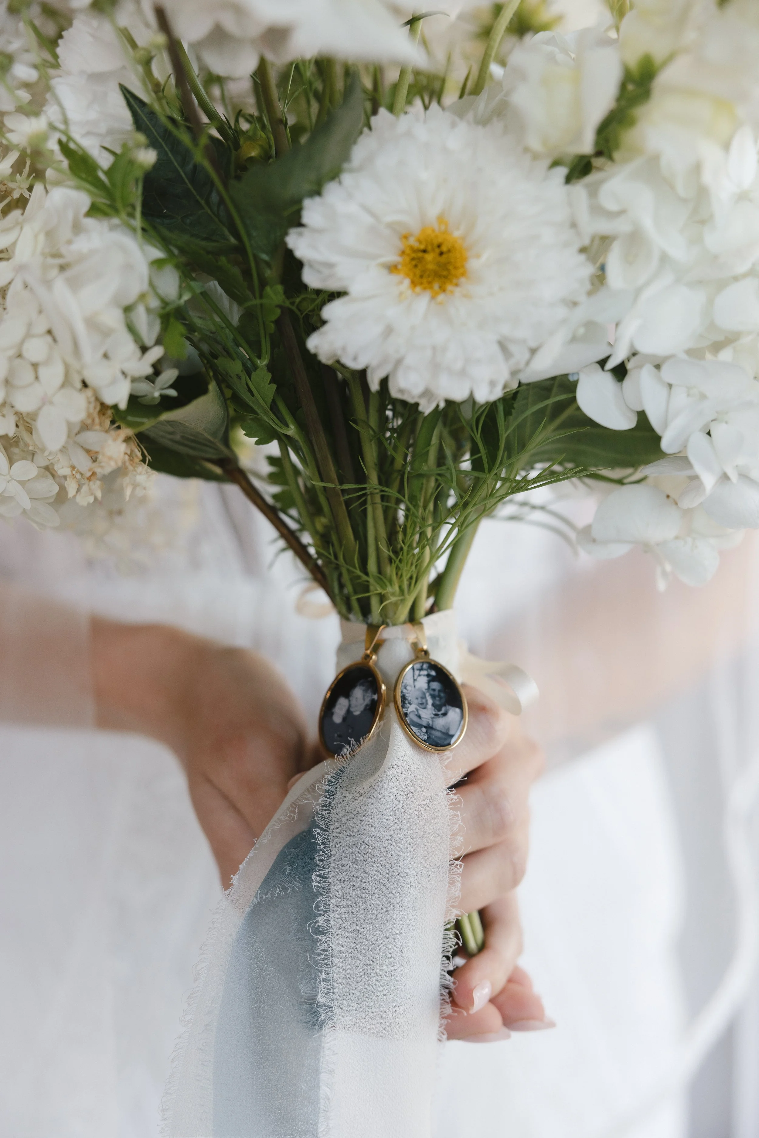 Person holding a bouquet of white flowers with a ribbon and two photo charms.
