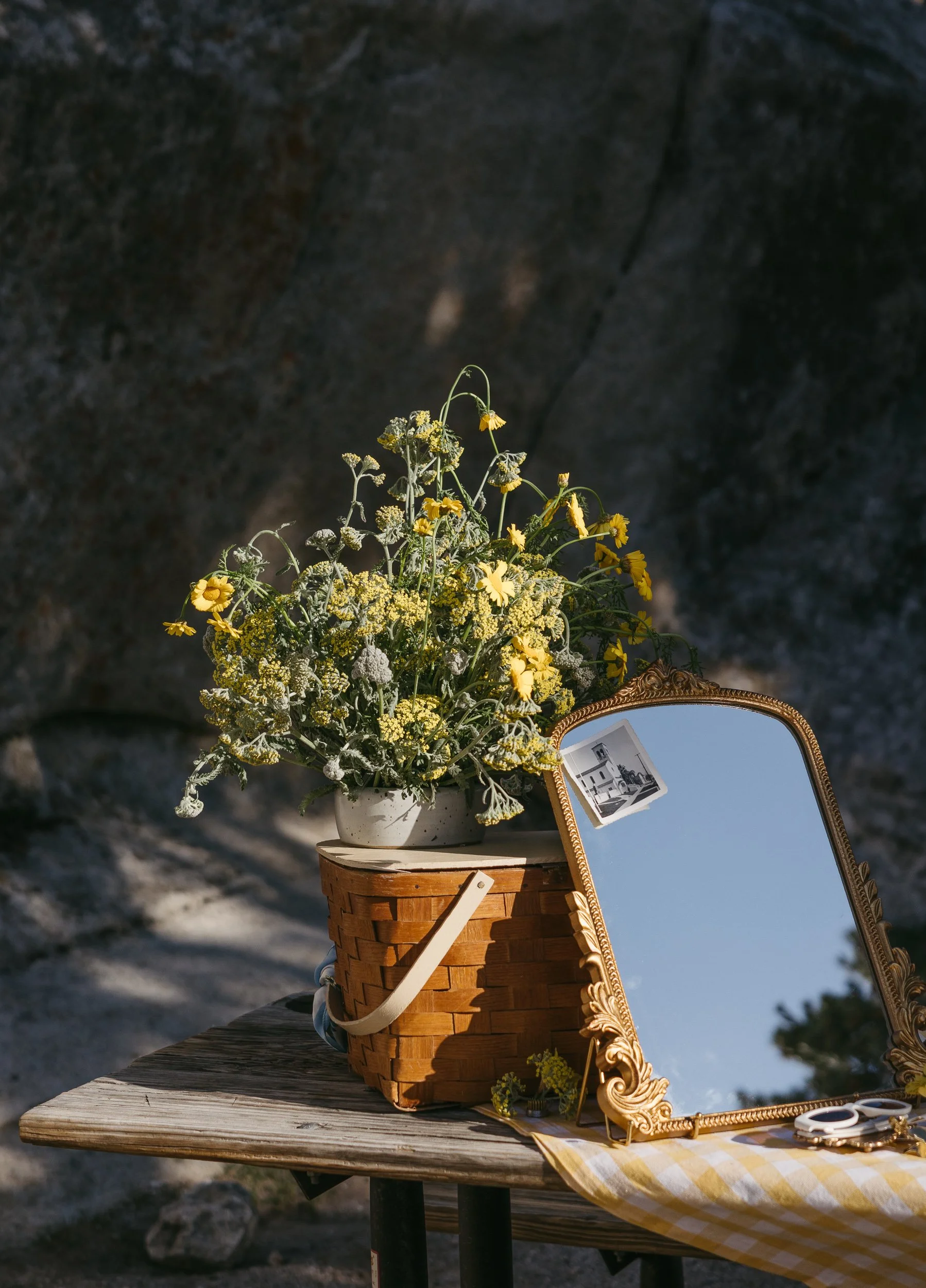 A wooden table with a basket holding a potted yellow and white flowers, a decorative mirror with a black and white photo clipped to it, and some small items on a yellow and white checkered cloth, outdoors against a rocky background.