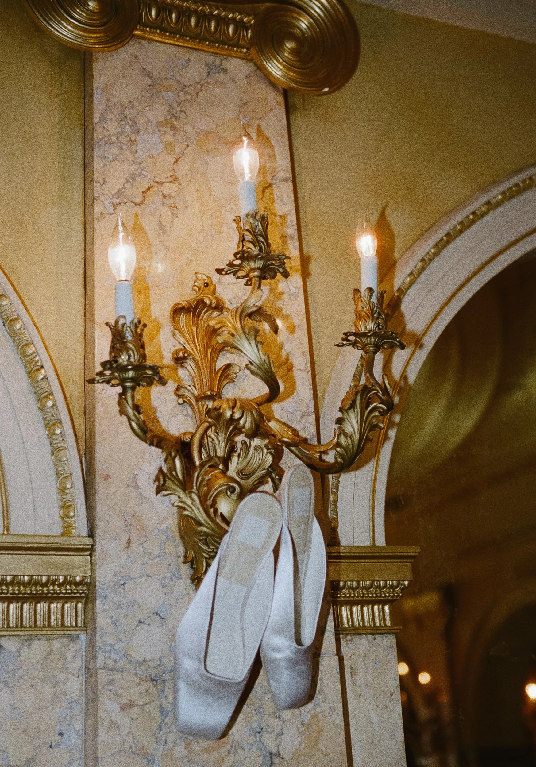 Ornate gold-colored antique wall sconce with three candle-shaped lights, decorated with leaves and scrolls, mounted on a marble wall with gold accents, with a pair of ballet slippers hanging from the bottom.