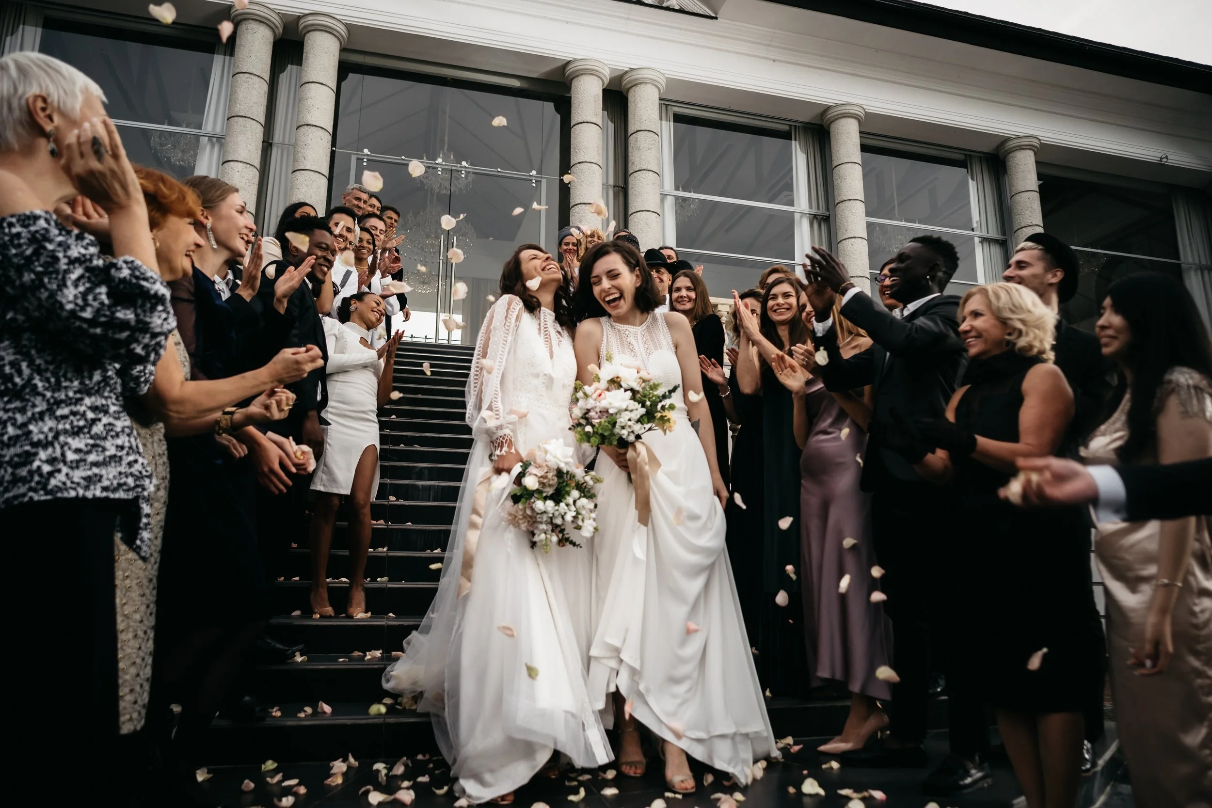 Two brides in white wedding dresses walk down the stairs hand in hand, smiling, surrounded by celebrating wedding guests throwing flower petals outside a building with large glass windows and stone columns.