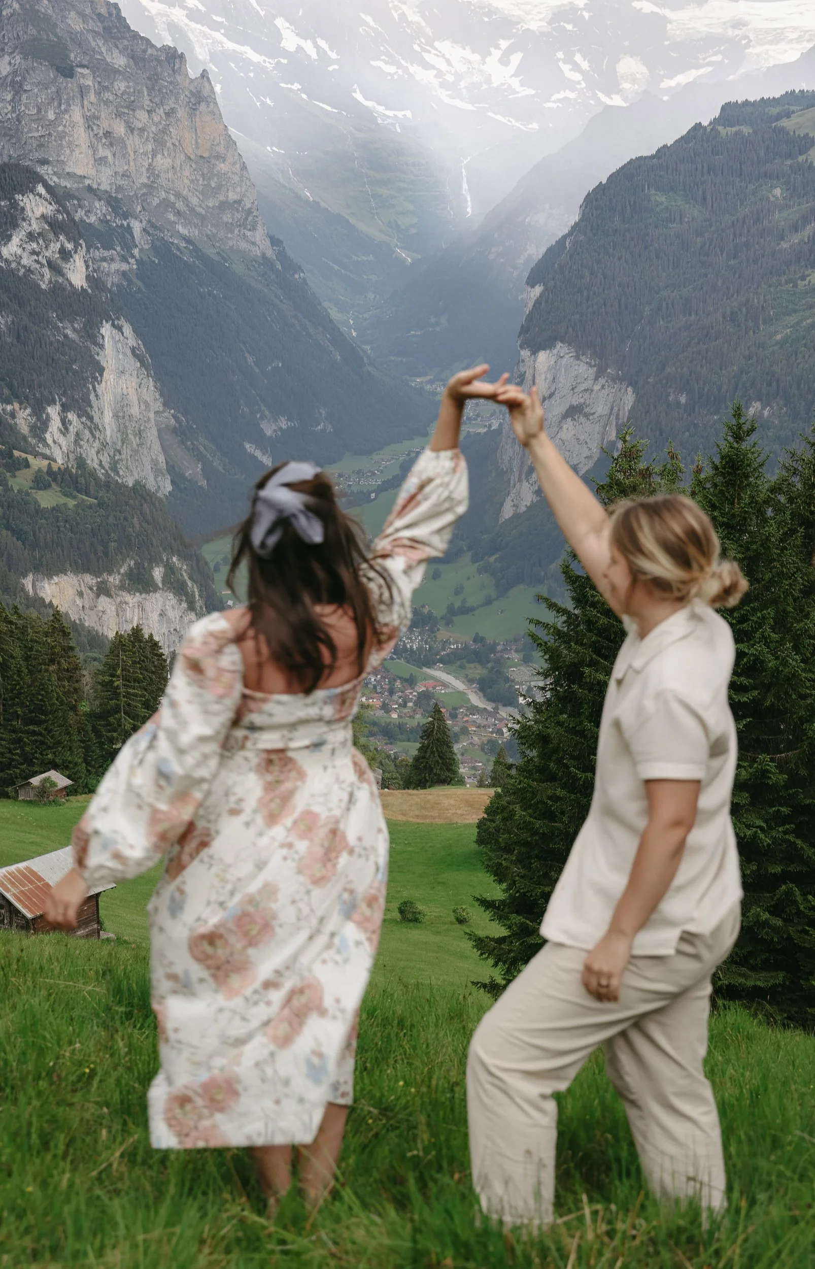 Two women dancing on a grassy hill with a mountainous valley in the background, surrounded by trees.